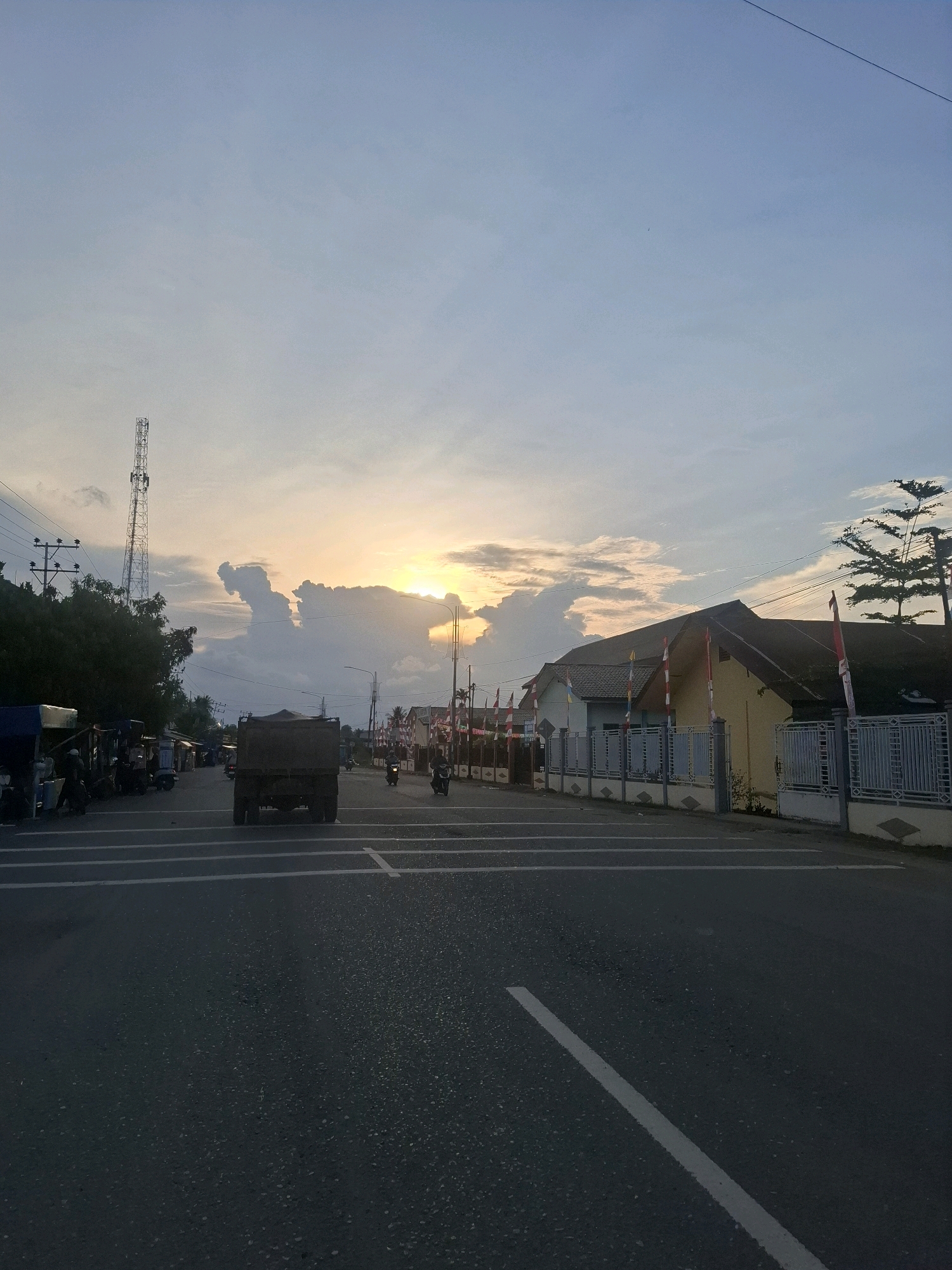 The image depicts a street scene at dusk in Rhieng Blang, Indonesia. A truck is driving down the road, which is lined with houses and shops on the right side. Indonesian flags are visible on the houses. On the left side of the road, there are several motorbikes parked in front of shops. The sky is a pale blue with some clouds, and the sun is setting behind them, casting a warm glow. A tall telecommunications tower is visible in the distance. The road has white lane markings. The overall impression is of a quiet evening in a residential and commercial area.