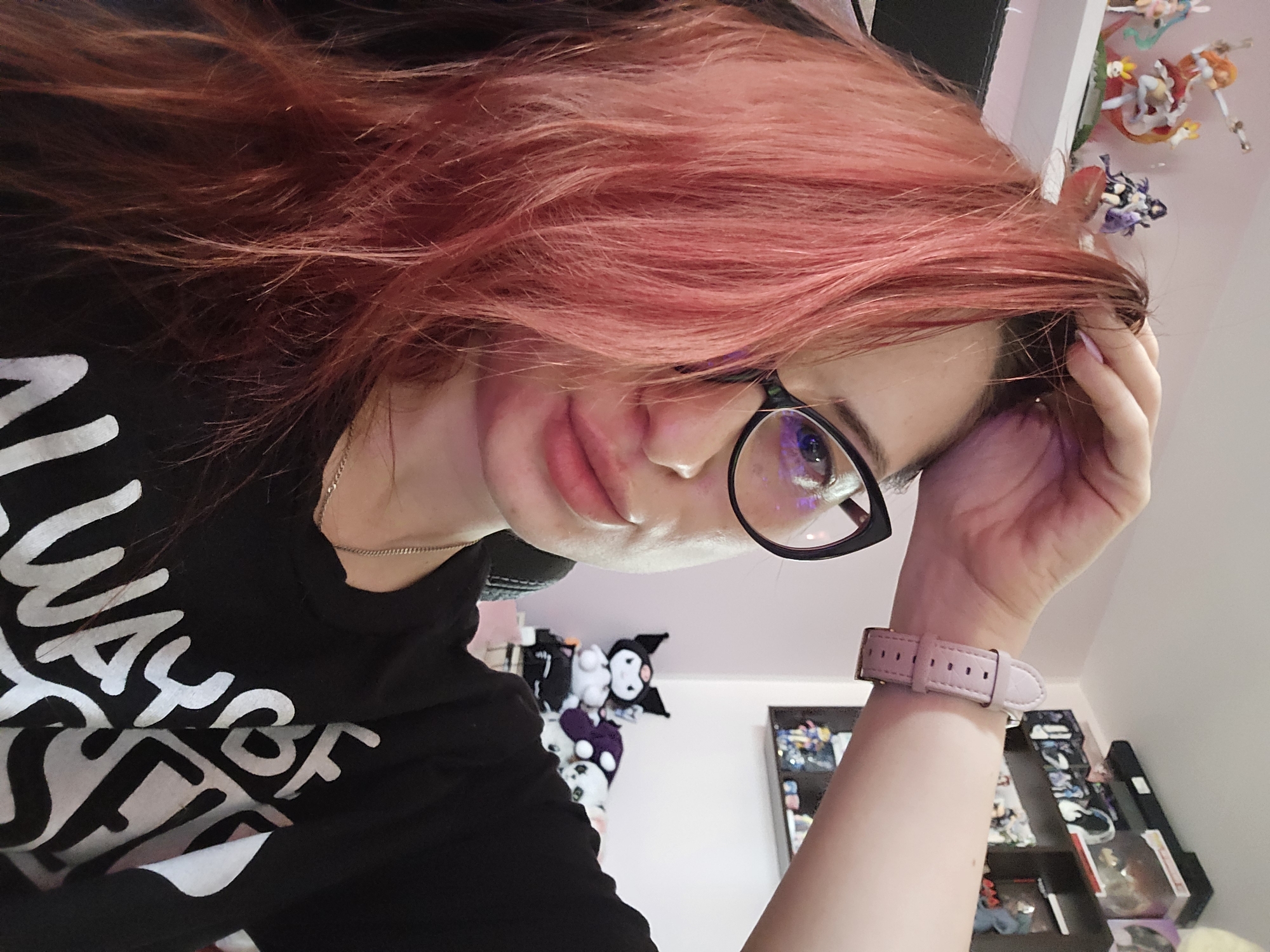 This is a selfie of a young woman with dyed pink hair. She is wearing a black t-shirt with white text, glasses, and a light pink watch. Her right hand is in her hair, and she is looking at the camera. The background shows shelves filled with anime figurines and stuffed animals. The lighting suggests it is daytime indoors. The overall mood is casual and personal. The text on her shirt is partially visible and appears to say "ALWAYS BE". The location is specified as Burgas, Bulgaria.