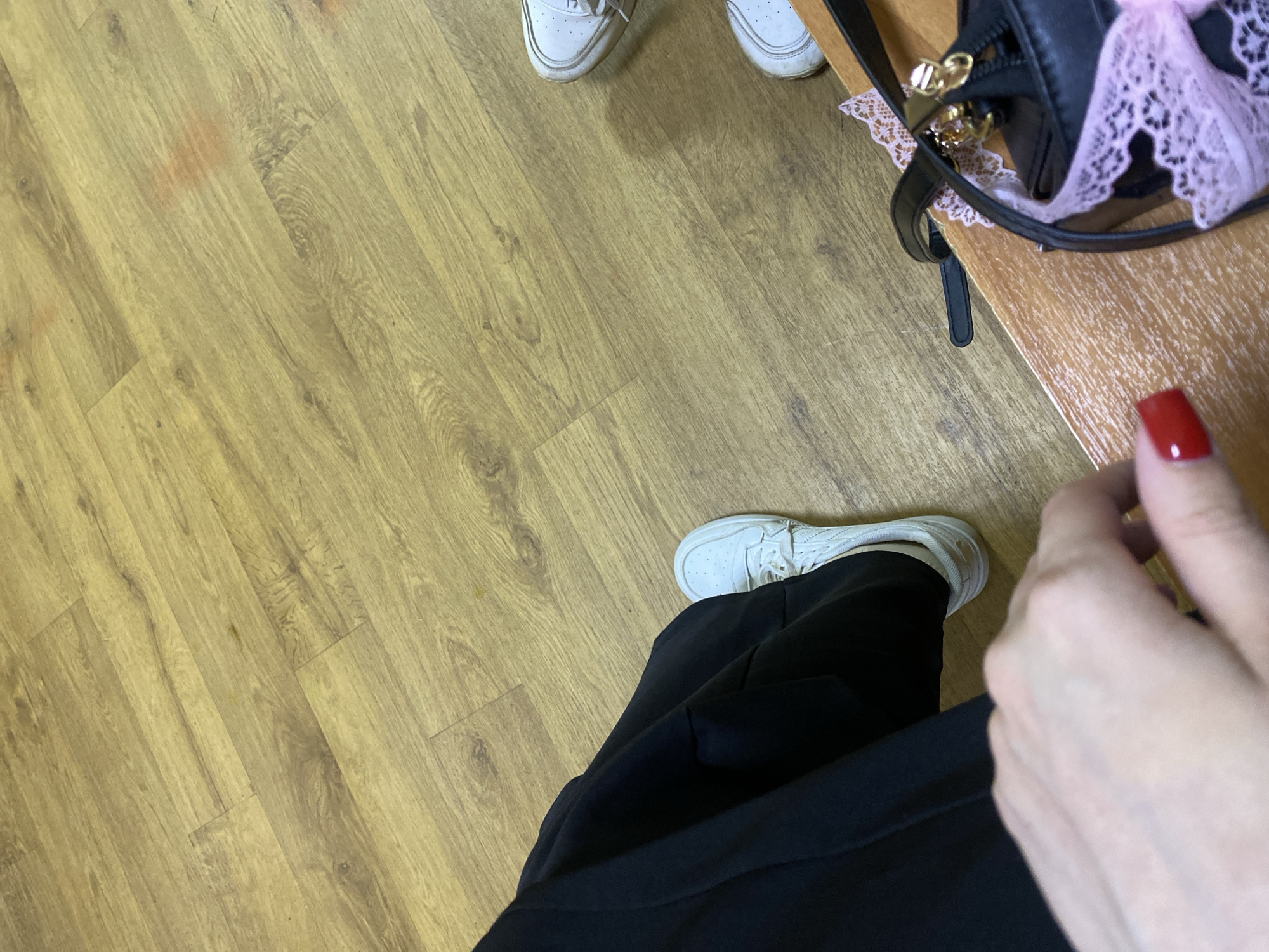 The image captures an indoor scene from a low-angle perspective, primarily showing a wooden floor, a person's lower body, and a handbag. In the immediate foreground, a person wears black pants and white sneakers. Their right hand, with bright red nail polish, is visible on the right side of the frame. Further back, another pair of white sneakers is seen, indicating a second person present.
A black handbag with gold metal hardware is placed on a light brown wooden surface, possibly a table or bench, located on the upper right side of the image. A delicate pink lace trim or accessory is attached to the handbag, adding a touch of color. The floor is covered with light-colored, plank-style wood or laminate flooring, displaying wood grain patterns. Several faint, reddish-brown smudges are noticeable on the floor.
The activity appears to be stationary, with individuals either seated or standing in a casual setting. The overall ambiance is relaxed and informal. No specific time of day or location cues are discernible beyond being an indoor space. There is no visible text within the image.