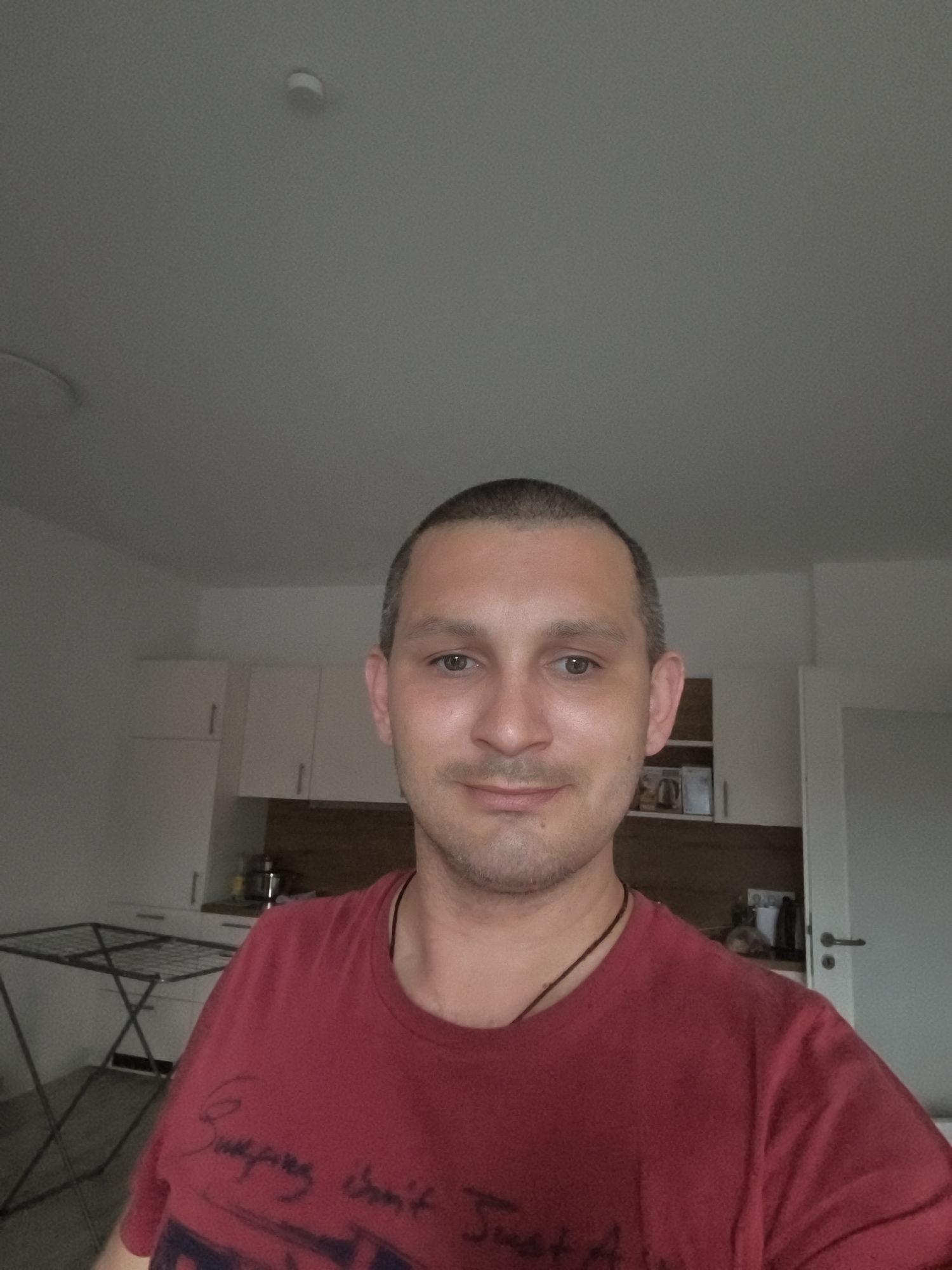 The image is a selfie of a man with short brown hair and light brown eyes, wearing a red t-shirt. He is indoors, in what appears to be a kitchen or living area. In the background, there are white cabinets, a wooden countertop, and a drying rack. A door with a silver handle is visible on the right side of the frame. The man is looking directly at the camera and smiling. There is no visible indication of the specific location within Wolgast, Germany.