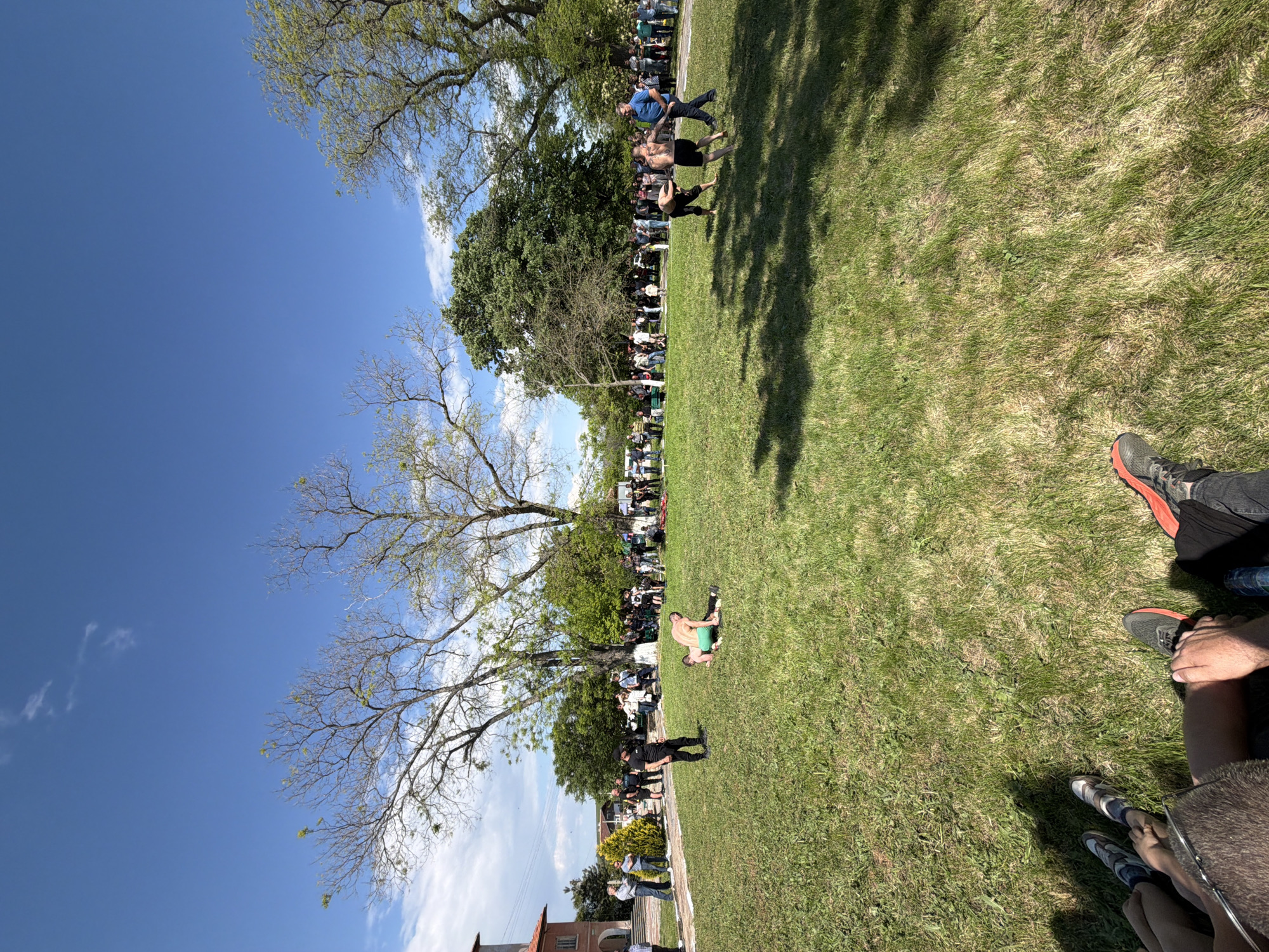 The image depicts an outdoor scene, possibly in a park or grassy area in the city of Dragomir, Bulgaria. People are gathered, appearing to observe something taking place on the grass, where at least one individual is lying down, wearing green clothing. A line of spectators can be seen in the background, along the edge of the grass. Trees provide shade and frame the scene, with a clear blue sky visible above. The buildings appear at the very edge of the image, suggesting the edge of the populated area or town.