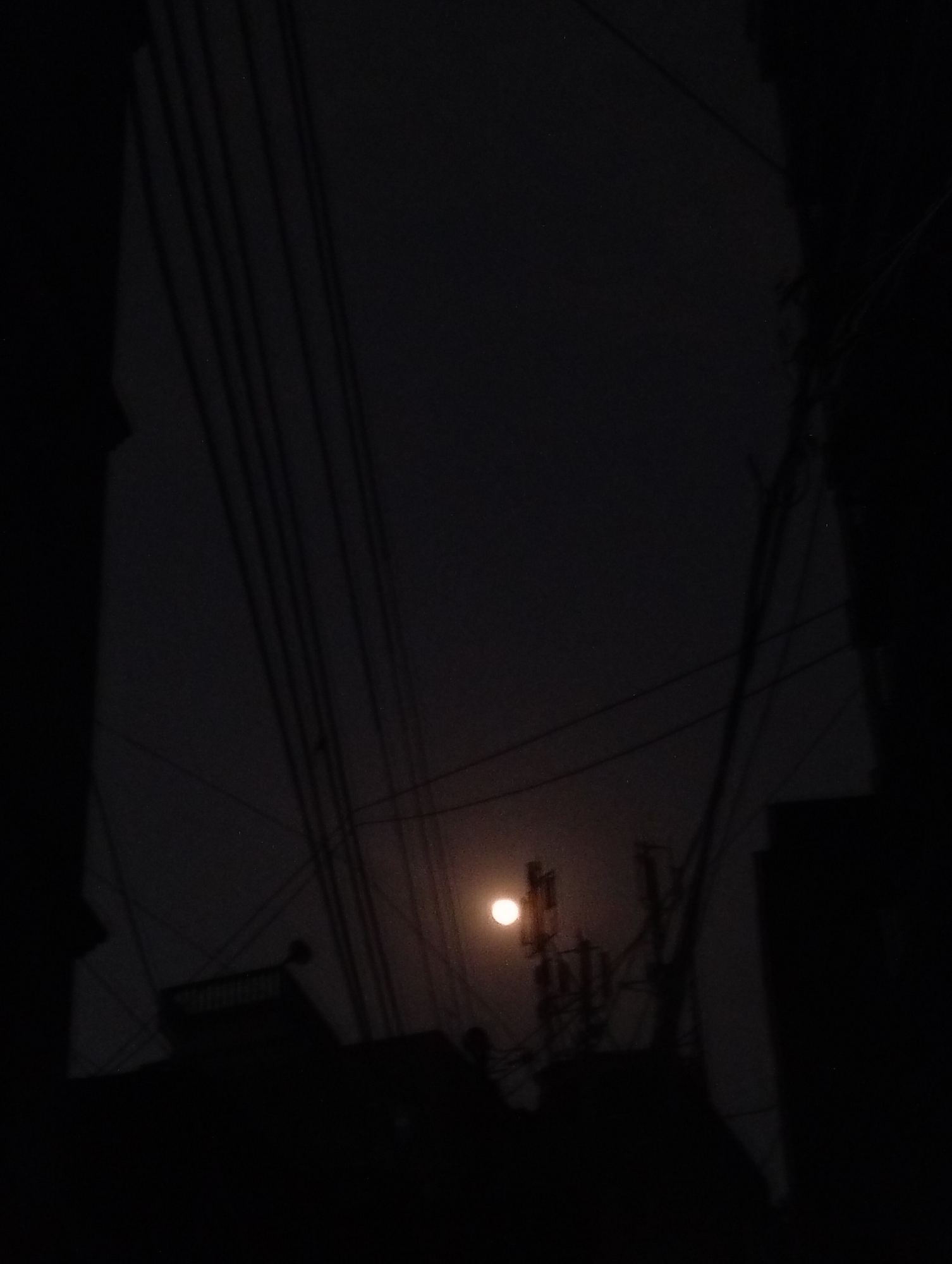 The photograph, likely taken in Dhaka, Bangladesh, depicts a night scene dominated by darkness. The light source is a dim moon or street lamp positioned behind a telecommunications tower and a web of power lines. The silhouettes of buildings and surrounding structures are visible in the foreground, framed by what appear to be the edges of a window or doorway. The scene is devoid of people and lacks discernible movement or interaction.