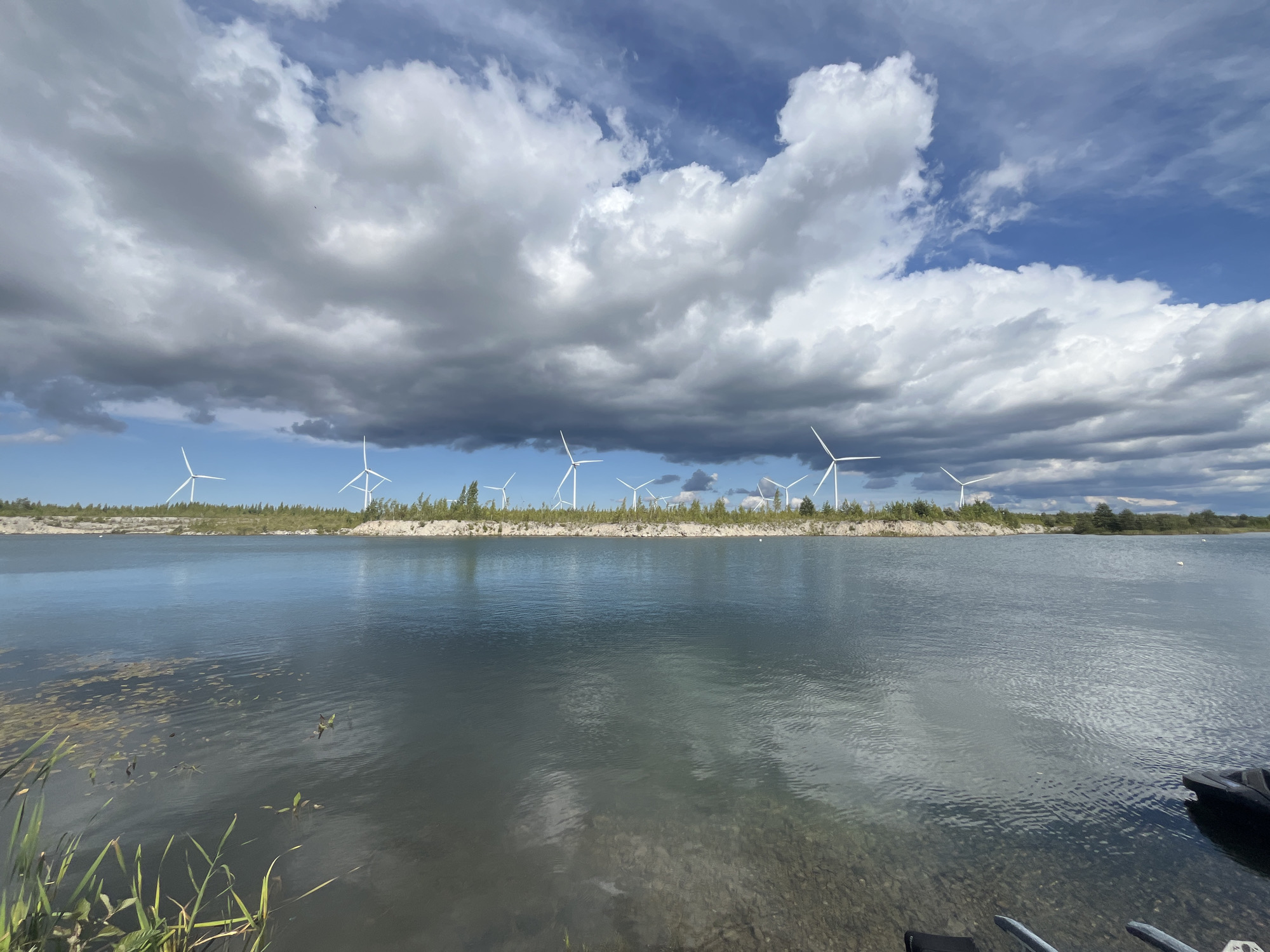The image depicts a body of water with a shoreline in the background. The shoreline is populated with numerous wind turbines, suggesting a wind farm. The sky above is partially cloudy with patches of blue sky visible. Vegetation, including trees and bushes, grows along the shoreline. In the foreground, the water is calm with some aquatic plants and debris visible. The bottom of the lake is visible in some areas due to the clear water. A portion of a black object, possibly a watercraft, is visible in the lower right corner of the image. The scene appears to be located in a natural or semi-natural environment. The image is a wide shot, capturing a broad view of the landscape.