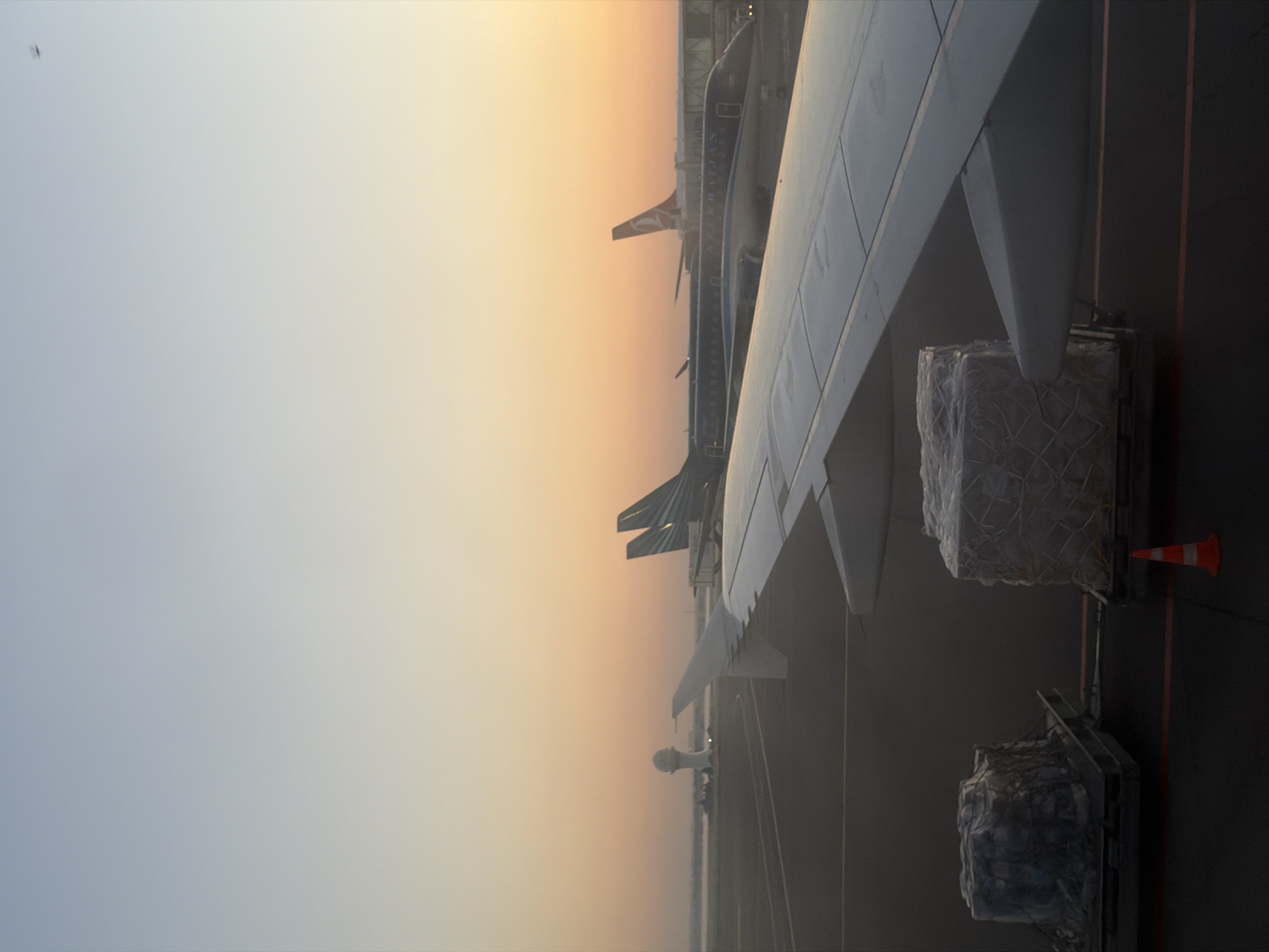 The image depicts an airport tarmac at dawn, with a clear sky transitioning from pale blue to a soft orange hue. In the foreground, the wing of an airplane is visible, casting a shadow on the tarmac. To the left, two cargo containers, wrapped in plastic and netting, are secured on palettes. To the right, an orange traffic cone is placed on the tarmac. In the middle ground, several aircraft are parked. One airplane, appearing to be an Embraer jet, is dark blue with "AZERBAIJAN" written on its fuselage. Further in the background, another airplane with a red tail fin, possibly Turkish Airlines, is visible. In the far distance, a radar tower stands prominently on the left side of the frame, with a hint of a body of water behind it. The overall scene suggests readiness for flight operations as the day begins.