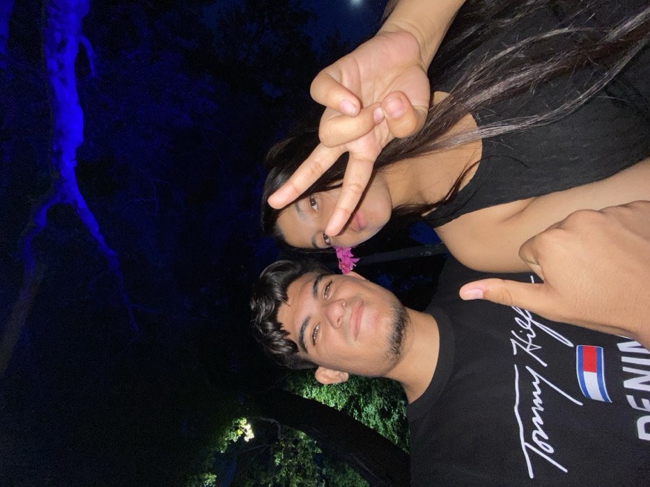 A young man and woman pose for a selfie at night. The man wears a black t-shirt with the Tommy Hilfiger logo. A pink flower is tucked behind his ear. The woman, positioned behind him, makes a peace sign with her fingers. A tree illuminated with blue light is visible in the background. The photo is rotated slightly, giving it a tilted perspective.