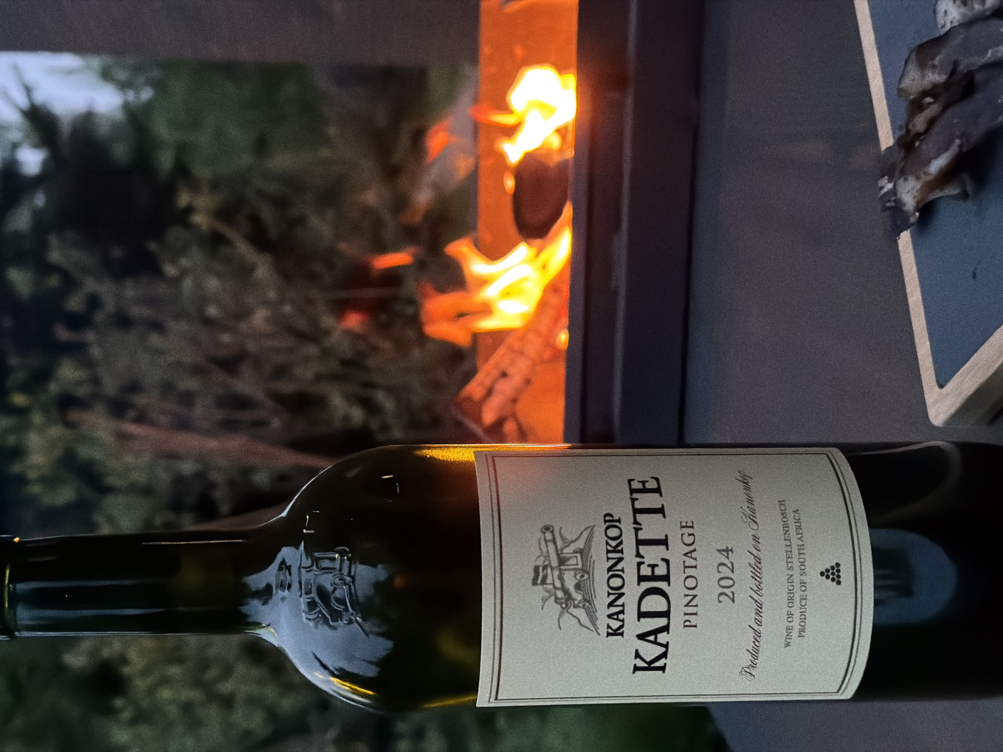 The image depicts a bottle of Kanonkop Kadette Pinotage 2024 lying horizontally in front of a fireplace. The label on the bottle is clearly visible, indicating it's a wine from Stellenbosch, South Africa. In the background, flames are burning in the fireplace. The setting appears to be outdoors, with greenery blurred in the background. The city of Zimyonki (Zimenki, Zimenka), Russia, is not identifiable or apparent in this image.