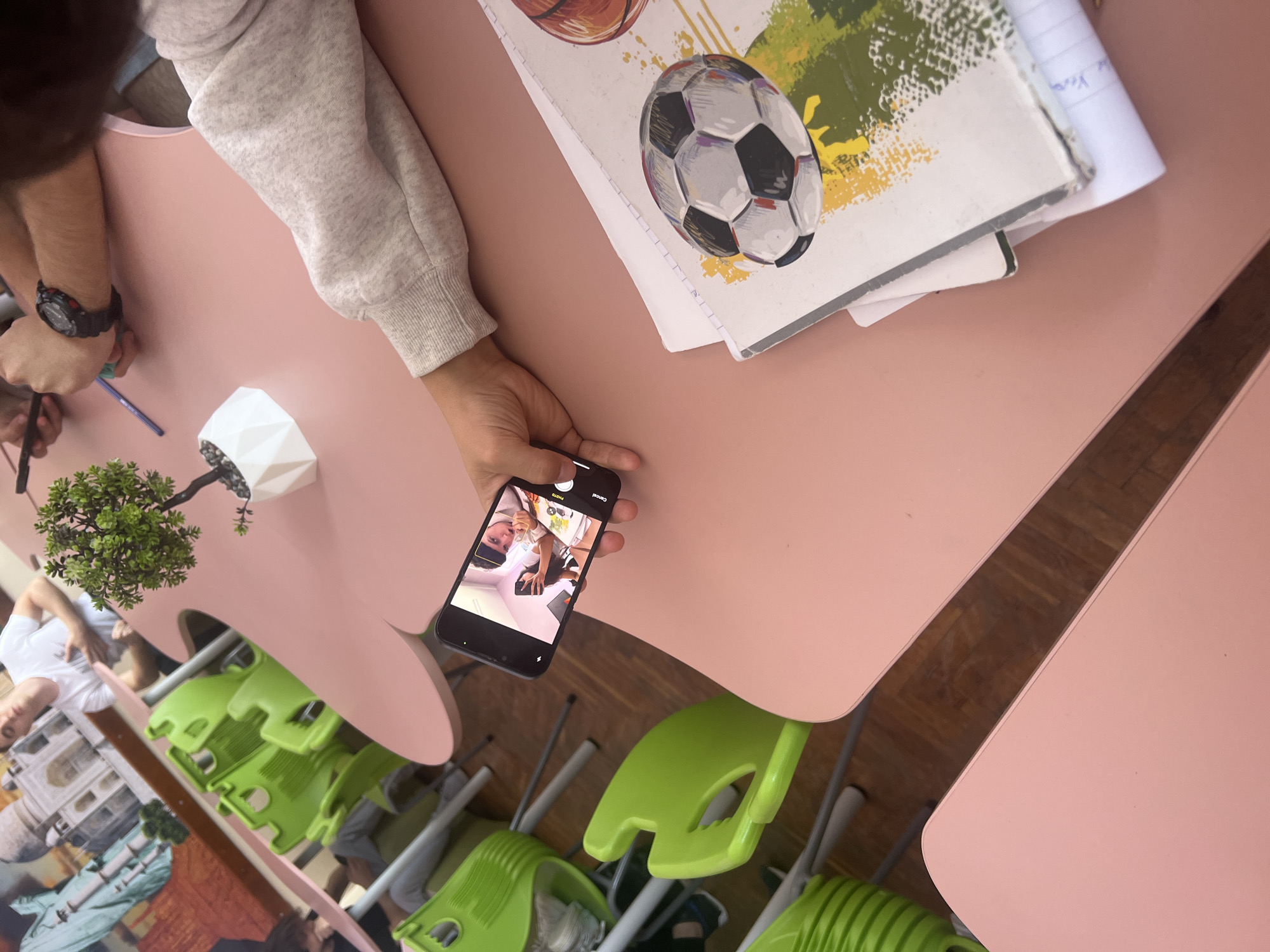The image presents an overhead, slightly tilted view of a classroom or study area featuring light pink, irregularly shaped tables and bright green chairs.
In the foreground, a person's arm, clad in a light grey sweatshirt, holds a black smartphone. The phone's screen displays a reflection of at least two individuals, one of whom, wearing a white shirt and glasses, appears to be looking towards the phone. Visible text on the phone screen indicates "Photo" and "Cancel," suggesting the camera application is active.
To the left, another person's hands are visible on the pink table, with one wrist wearing a black digital watch. A blue pen or pencil is held in their fingers, resting near the table's edge. A small artificial plant in a white geometric pot sits between the two individuals.
Further on the table, two notebooks are stacked. The top notebook features colorful illustrations of a soccer ball and a basketball. On the notebook beneath it, faint handwritten text, possibly "Yoo," is discernible. The floor beneath the furniture is composed of wooden planks. The chairs are partially stacked or arranged in a way common in classrooms. The scene depicts a casual, indoor setting, likely a school or university, where individuals are engaged, possibly in studying or social interaction facilitated by the smartphone.