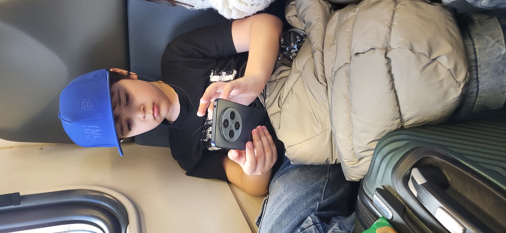 A young boy wearing a blue baseball cap and a black t-shirt sits on a train, engrossed in his smartphone. He is using earbuds connected to the phone. The boy is covered by a beige puffer jacket and is wearing blue jeans. In front of him, a dark green suitcase with silver accents is visible. The background shows the interior of the train, with a window on the left and a dark gray seat.