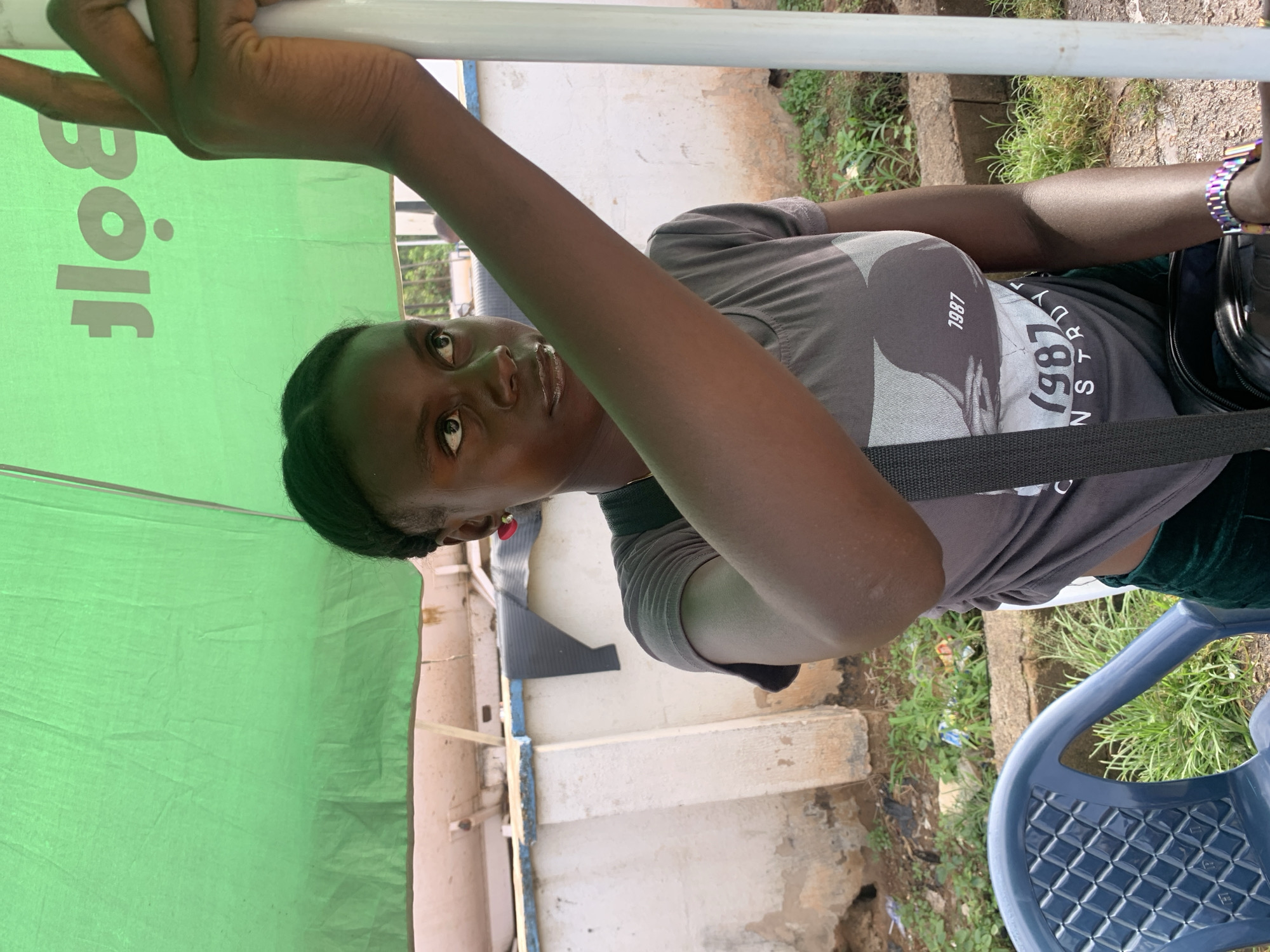 A young Black woman, with dark hair pulled back, is seated outdoors. She is wearing a grey t-shirt with a graphic print and the year "1987" on it, and dark green pants. A black strap from a bag is visible across her chest. She has a colorful watch on her left wrist. She is leaning against a white pole and looking upwards and to her left. A bright green umbrella or canopy is visible above and behind her, with a logo that appears to read "Bolf" in white lettering. In the foreground, a blue plastic chair is partially visible. The background shows a rough, textured wall, some overgrown vegetation, and debris on the ground. The scene suggests an outdoor setting, possibly a street-side vendor or a casual waiting area in Jos, Nigeria.