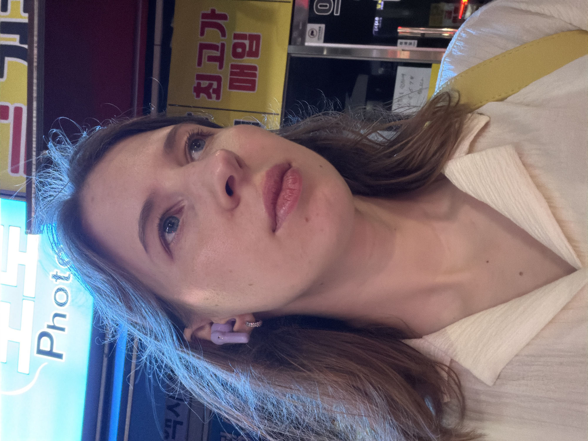 A light-skinned woman with light brown hair is pictured from the chest up, facing to the right. She is wearing a light-colored collared shirt and a yellow strap across her shoulder. She has on purple earbuds and a small hoop earring in her visible ear. The background appears to be an outdoor urban setting at night, with illuminated signs. One sign in the upper left corner displays the word "Photo" in large, bright blue letters against a blue background. Another sign in the upper right features Korean text in red lettering on a yellow background, including "최고가" and "매입". The overall impression is a selfie taken in a city.