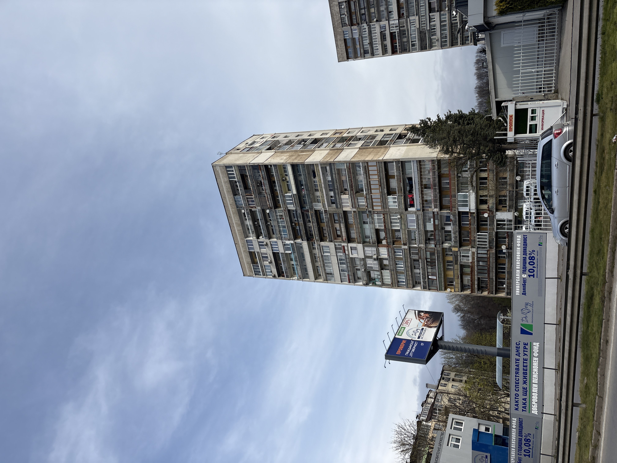 This image depicts a typical urban scene in Sofia, Bulgaria, featuring a Soviet-era residential high-rise building. The building is constructed of concrete and has numerous balconies and windows, indicative of its residential function. A second building, partially visible, is of similar construction. In the foreground, a large billboard advertises pension funds and insurance services, written in Bulgarian Cyrillic. A gray car is parked near a checkpoint booth, and some trees are visible. The sky is overcast, suggesting a cloudy day. The environment is urban, with a blend of residential buildings, advertising infrastructure, and street-level elements.
