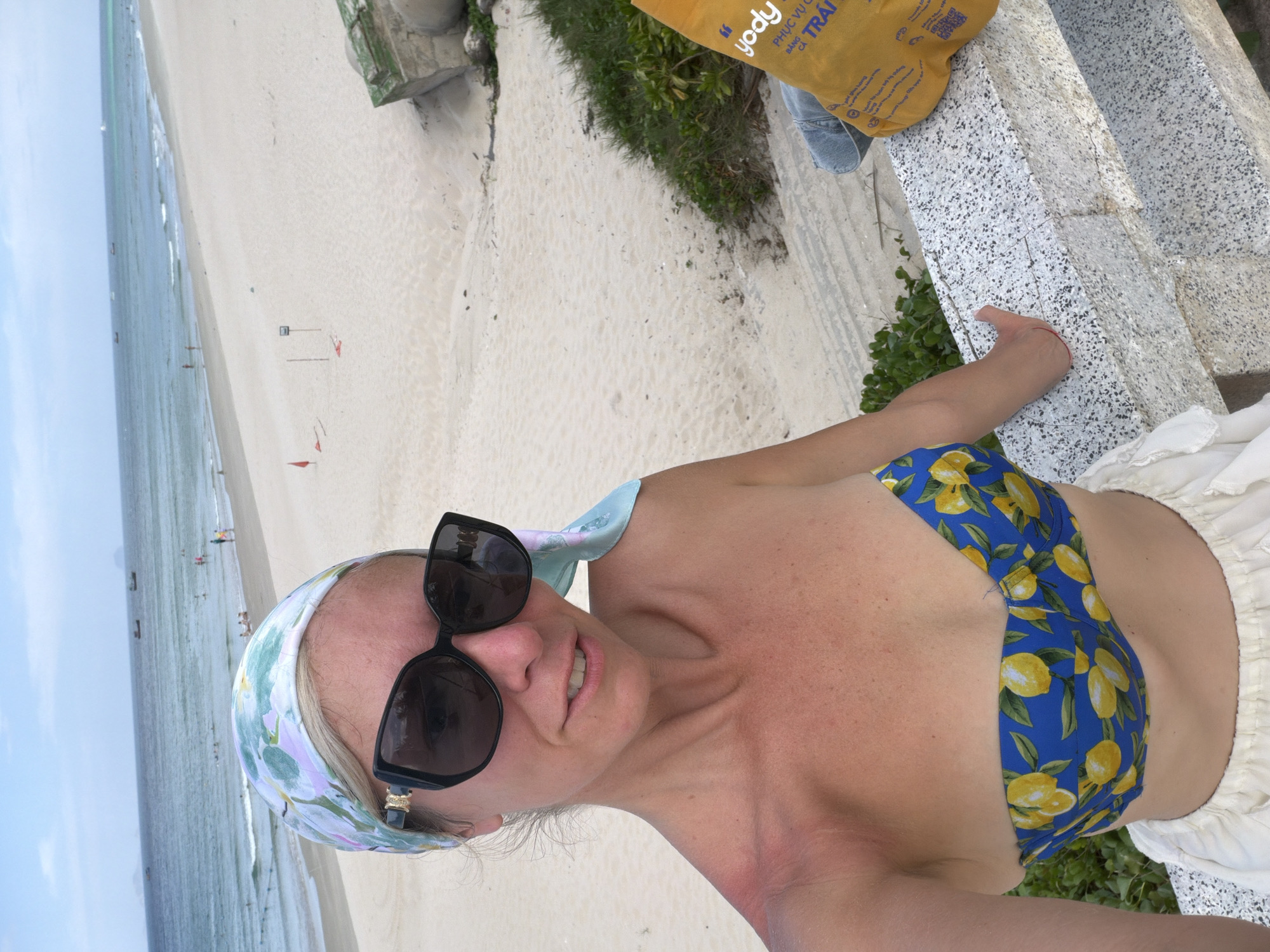 A woman with blonde hair, wearing a floral headscarf and sunglasses, poses for a selfie on a beach in Cam Đức, Vietnam. She is wearing a blue bikini top with a lemon print and a white skirt. In the background, there is a sandy beach, the ocean, and a cloudy sky. There are a few boats in the water and some people on the beach. To the right, there is a yellow bag with Vietnamese writing on it, and some greenery. A concrete structure is visible in the upper right corner. The woman has a sunburn on her left shoulder.
