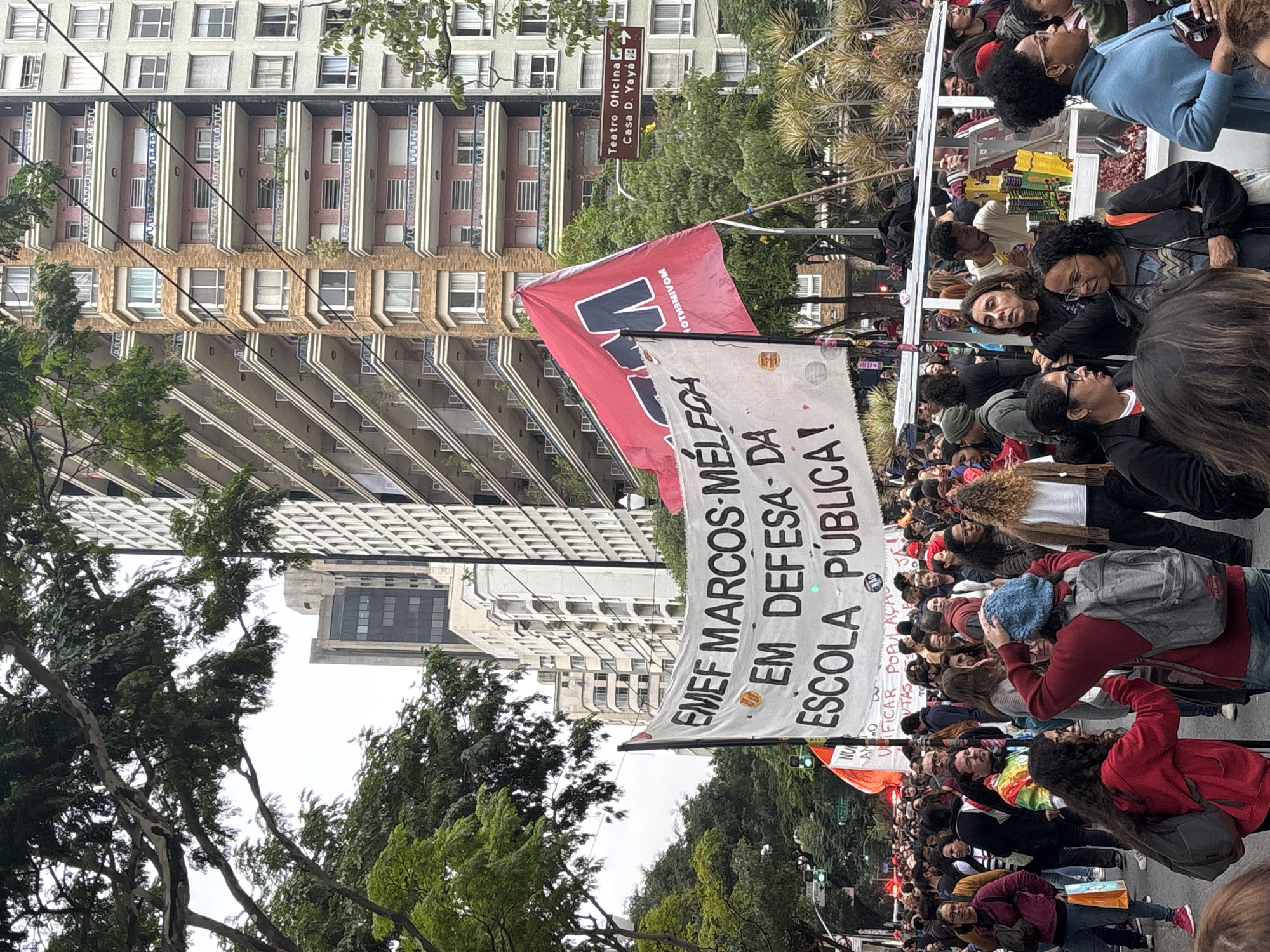 The image captures a demonstration in São Paulo, Brazil, likely focused on education. A large group of people is gathered, carrying banners that read "EMEF MARCOS MEL FOH EM DEFESA DA ESCOLA PÚBLICA!" (referencing EMEF Marcos Melfoh and advocating for public schools). A red flag with black lettering is also visible.
The demonstration is taking place on a city street, with tall buildings lining the background. A sign pointing to "Teatro Oficina" and "Casa D. Yaya" suggests a location near those landmarks. People are of varied ages and ethnicities. Some are looking directly at the camera, while others are interacting with each other.