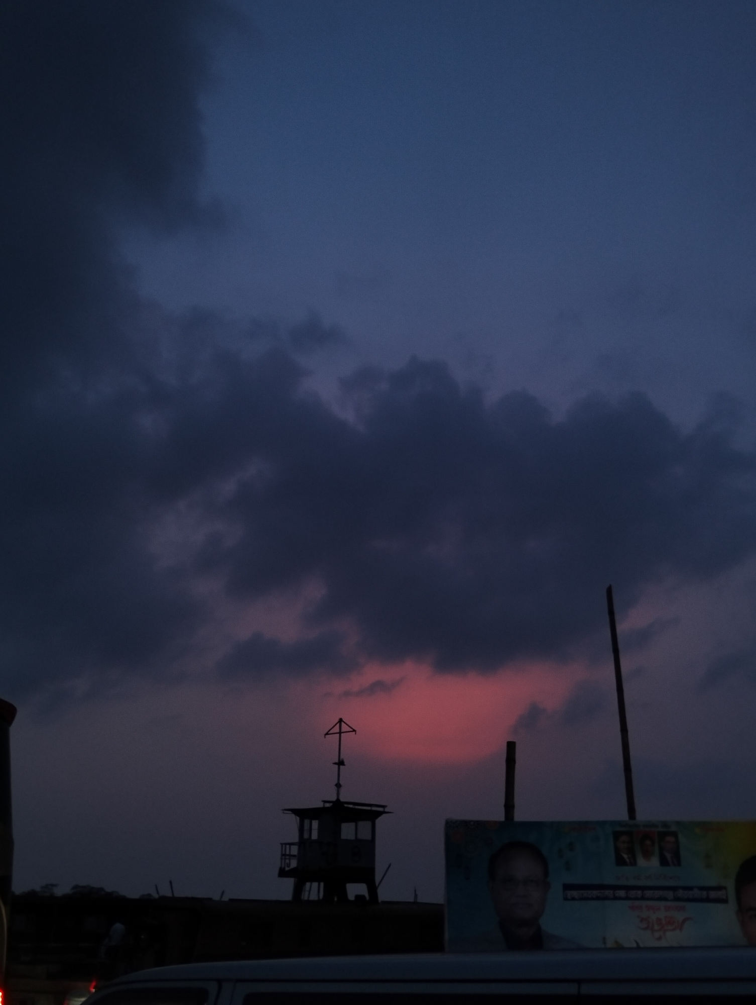 A low-angle, long shot captures a cityscape at dusk in Unknown, Unknown. In the foreground, the blurred top of a white van is visible. Behind it, a partially visible billboard displays portraits of three men. Adjacent to the billboard, a tall wooden pole stands vertically. To the left, a silhouetted guard tower is discernible. Above, dark clouds dominate the sky, illuminated by a faint pink glow on the horizon.