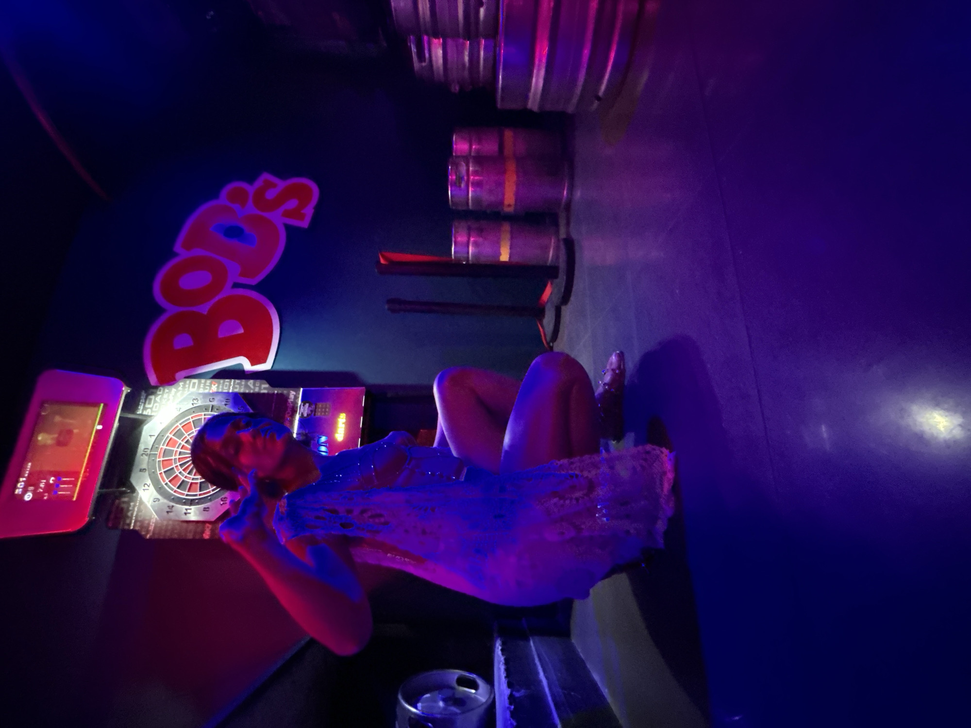 The image depicts a darkened interior space, identified as a bar or entertainment venue in Calella, Spain. The scene is heavily illuminated by red, blue, and purple neon lighting, creating a high-contrast, atmospheric environment.
A single young female is positioned in a low crouch in the foreground, facing generally towards the viewer. She has dark hair and is wearing a light-colored, possibly lace or crocheted, sheer top. A visible bra strap is present. Her bare legs are visible, and she is wearing studded sandals. Her right arm is raised, with her hand forming a "V" or peace sign. Her left hand appears to be holding a loose piece of fabric, possibly part of her attire.
In the background on the left, an electronic dartboard, displaying "501" on its screen, is prominent. The dartboard features a traditional numerical layout. To the right of the dartboard, a red neon sign spells out "Bob's" in a stylized, bubble font, with a blue circle within the "o". Several silver beer kegs are visible along the right wall, some stacked, and one partially visible on a raised platform to the far left. Two dark stanchions with red tops are positioned on the right side, between the dartboard and the main group of kegs. The floor is a dark, reflective surface, showing various light reflections from the neon signs. The walls are dark, serving as a backdrop for the illuminated elements.