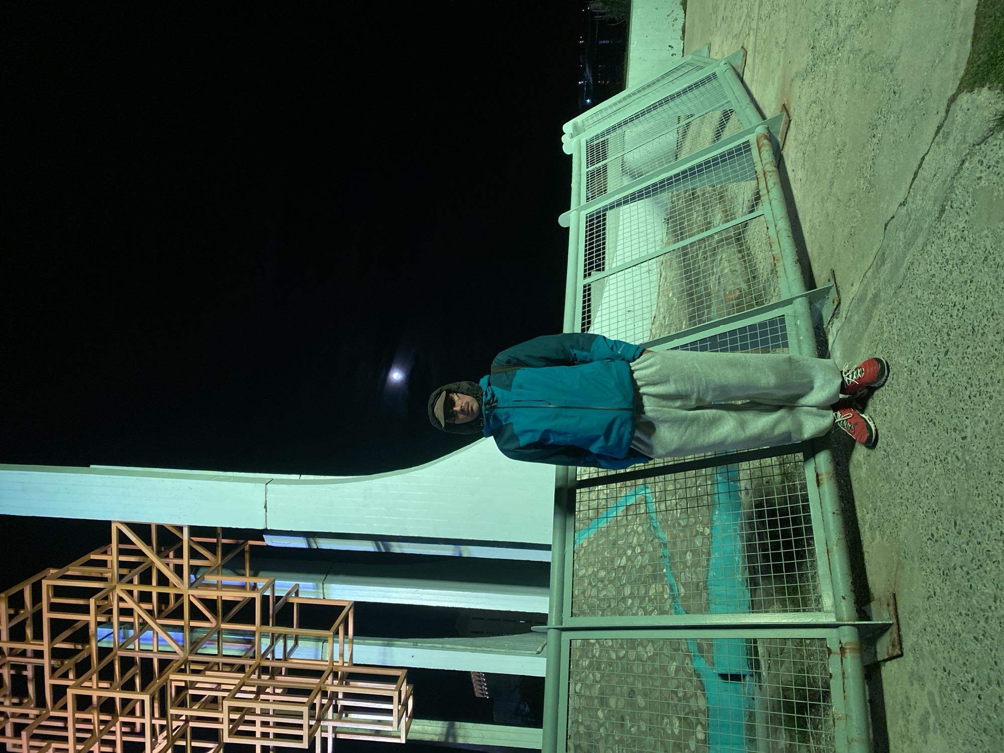 A person stands in front of a structure at night. The individual is wearing a blue jacket, grey sweatpants, red shoes, and a hooded sweatshirt pulled up, obscuring some of their face. They are positioned near a metal grid railing and concrete. To the left, a geometric structure made of wooden frames is illuminated. The background is dark, with a full moon visible. The scene appears to be located in a public space, likely in Ushuaia, Argentina, based on architectural styles and environmental conditions common in the region.