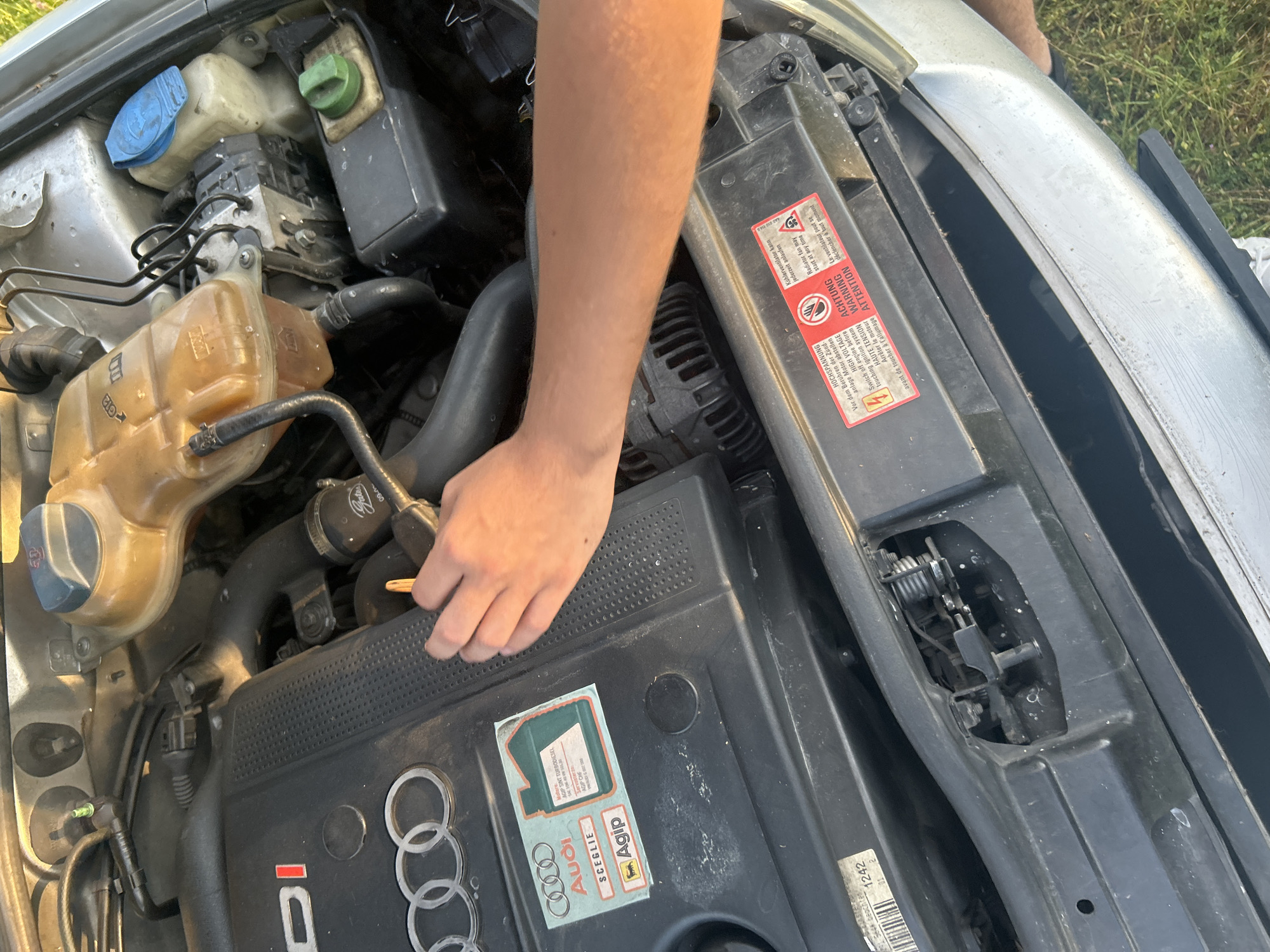 The image presents a close-up, overhead view of the open engine bay of a silver Audi vehicle, situated outdoors in the city of Lebed, Bulgaria. A person's tan forearm and right hand are prominently centered in the frame, resting on the engine's black plastic cover.
The engine bay contains various components:
- A black plastic engine cover features the silver Audi rings logo and the letters "DI," indicating a direct injection diesel engine.
- An "Audi SCEGLIE Agip" service sticker is affixed to the engine cover, depicting a green oil bottle and specifying "Agip SINT TURBODIESEL SAE 10W-40 VW 505 00" and "AGIP CHF VW 505 000."
- To the right of the engine cover, a translucent, yellowish plastic coolant expansion tank, marked with "G12" and "MAX/MIN" indicators, is connected by black hoses.
- Adjacent to the coolant tank, a white plastic brake fluid reservoir with a blue cap is visible, alongside an ABS pump module with multiple black lines.
- Below these components, a black reservoir with a green cap, likely for power steering fluid, is partially obscured.
- Various black rubber hoses, secured with metal clamps (one branded "Gates"), and electrical wiring are distributed throughout the engine compartment.
- Part of an alternator or similar pulley system is visible below the engine cover.
- On the front radiator support beam, a series of red and white warning labels are present. These multi-lingual warnings (German, English, French) caution about high voltage in the ignition system (with a lightning bolt symbol), advise switching off the engine before touching, and warn that the radiator fan may start automatically at any time (with a fan symbol).
The human hand is open, with fingers slightly spread, positioned over the textured surface of the engine cover near the Agip sticker. The car's silver front bumper or fender is visible at the bottom of the frame. Green grass is visible in the background, suggesting an outdoor setting under natural daylight.