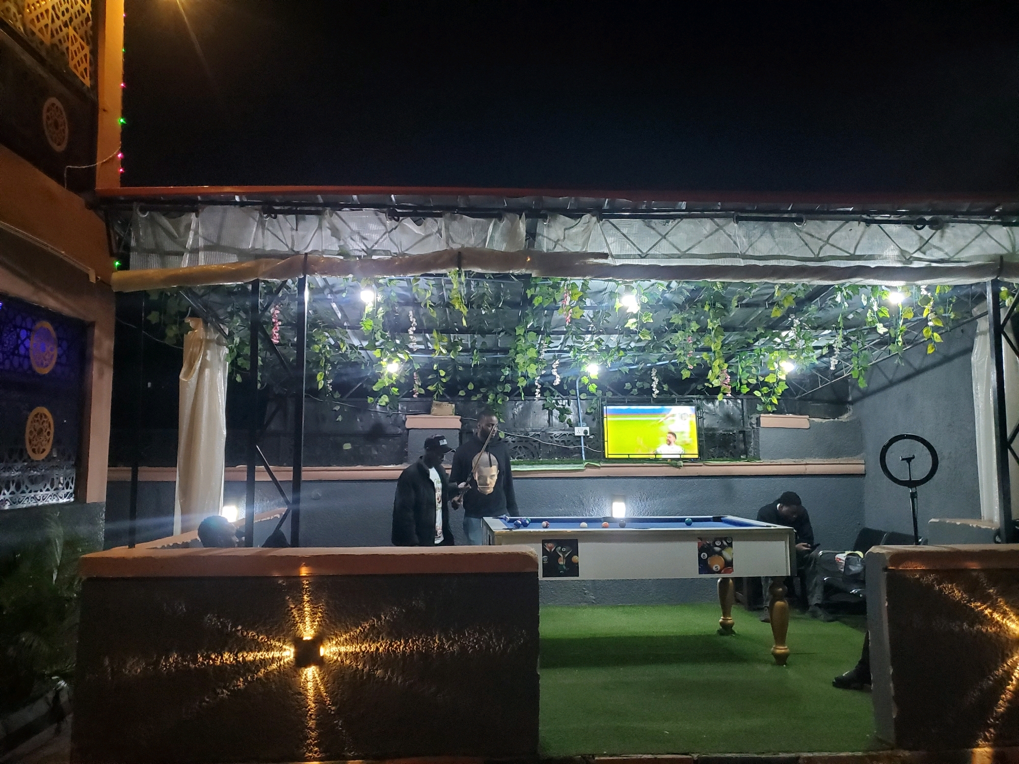 The image depicts an outdoor recreational space at night, likely in Jos, Nigeria. In the foreground, a decorative stone wall with embedded lights casts a starburst pattern onto the surrounding area. Beyond this, a green artificial turf covers the ground, leading to a pool table set up in the middle of the scene. The pool table has a blue felt surface and visible billiard balls.
Behind the pool table, several individuals are present. Two men are standing near the pool table, one wearing a black jacket and a black cap, the other in a black hooded sweatshirt with a print on the front. A television screen is mounted on the wall behind them, displaying an image. Further to the right, another person is visible, appearing to be using a smartphone. A ring light on a stand is also positioned in this area.
The space is partially covered by a structure with an overhang, from which artificial ivy and string lights hang, creating a decorative canopy. The background consists of a grey wall. The overall atmosphere suggests a social gathering or leisure activity taking place at an establishment or private residence.