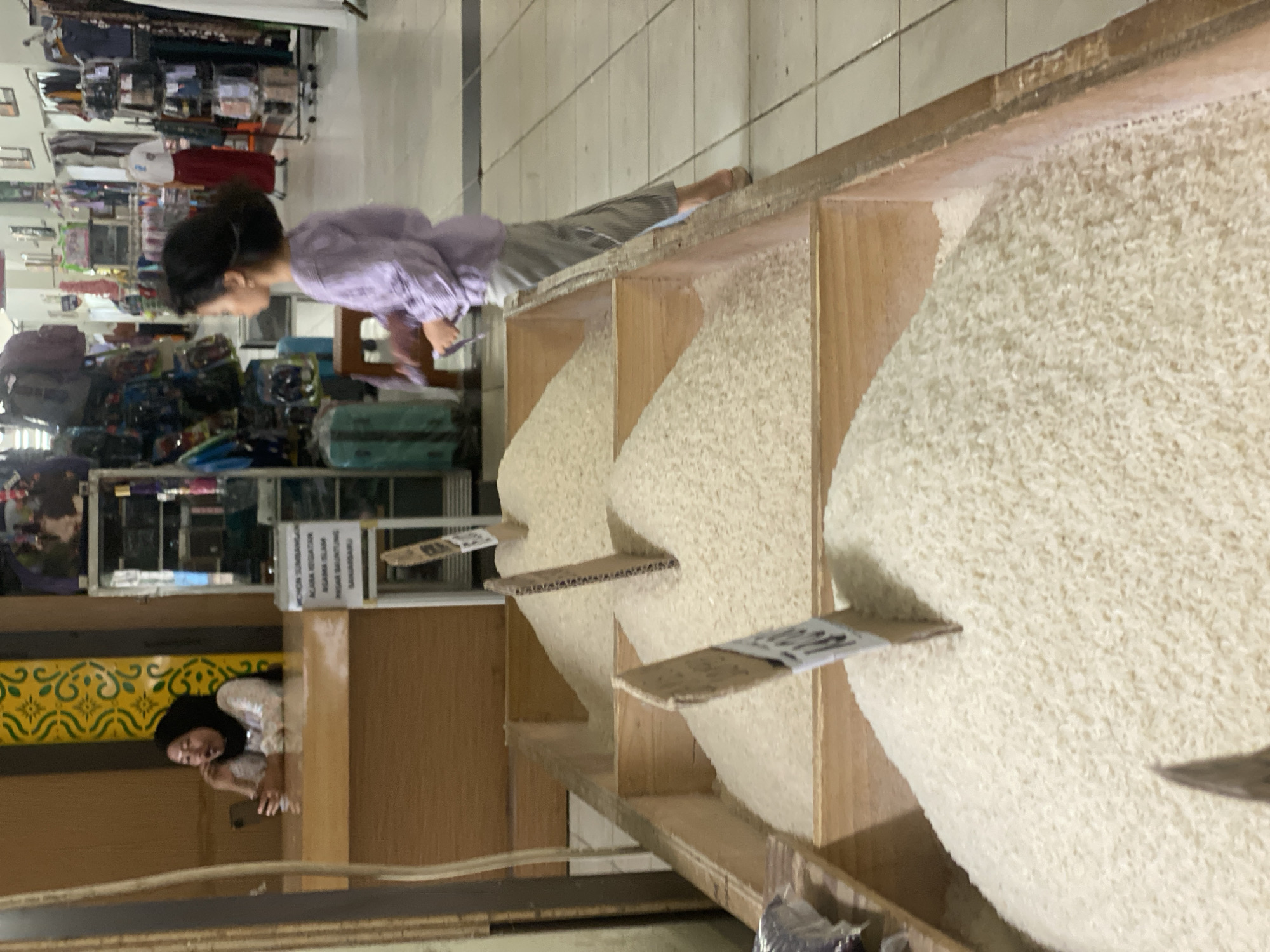 A young girl with dark hair, wearing a light purple striped shirt and gray striped pants, stands in a market in Banjarbaru, Indonesia, looking to her right. She holds a small, dark object in her right hand. In the foreground, three wooden crates are filled with large quantities of white rice. Cardboard signs are inserted into the rice in two of the crates. Behind the rice display, a woman wearing a hijab, a white top, and a dark patterned scarf sits at a counter, looking at a smartphone. The background shows shelves stocked with various goods, including bags and clothing. The market environment features tiled flooring and wooden structures.