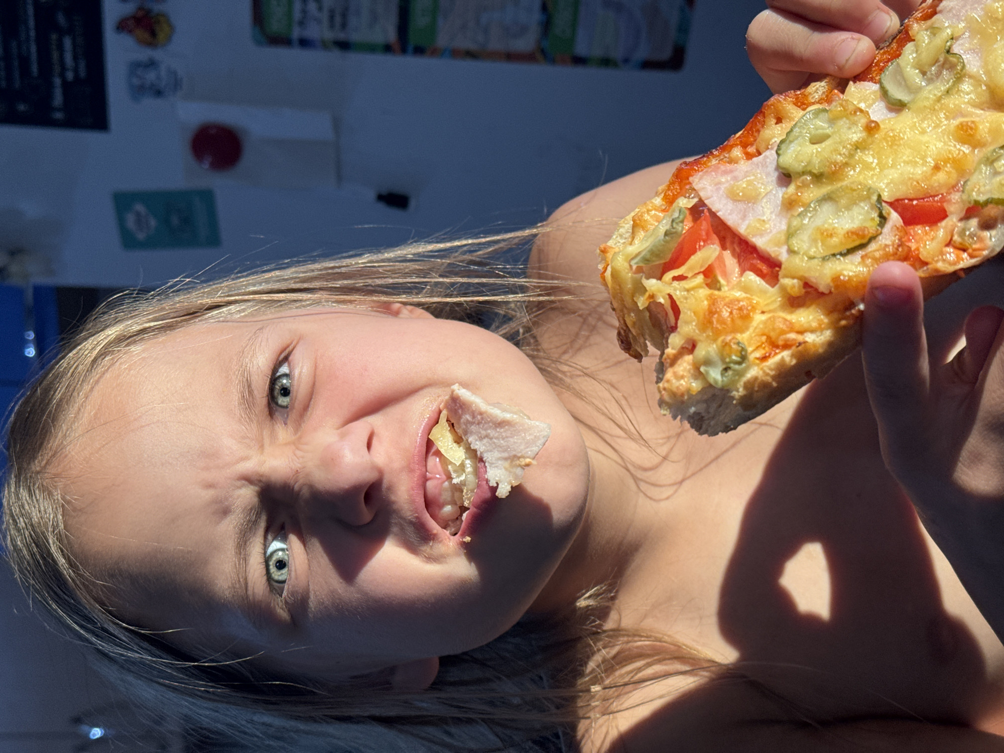 A young child is captured in a close-up, head-and-shoulders shot, eating a slice of pizza. The child's face is animated, with a scrunched-up expression, wide eyes, and a mouthful of food. They are holding the pizza slice with their right hand, bringing it closer to their mouth. The background is blurred, but appears to be an indoor setting with a blue wall and some items mounted on it, possibly in a kitchen. Sunlight casts a strong shadow on the child's chest and shoulder, suggesting the light source is from the left. There are no discernible elements that suggest a specific location within the city of Rusalevka, Russia. The image is focused on the child's interaction with the pizza.