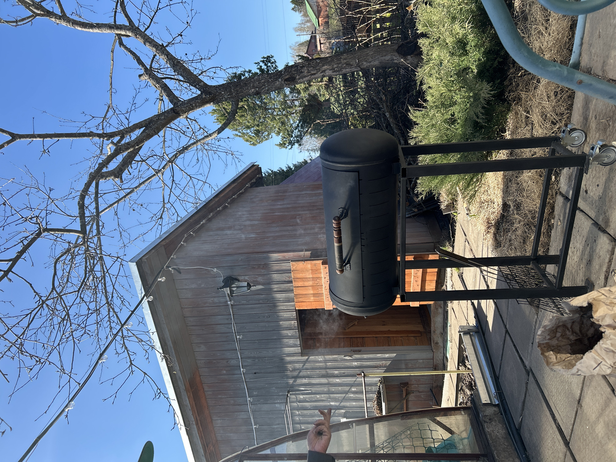 The image depicts an outdoor scene with a black cylindrical smoker situated on a black metal frame with wheels, resting on a paved surface next to a wooden building. The building has a window or opening through which smoke is visible. A bare tree branch is visible in the upper left corner against a clear blue sky. Part of a greenhouse is seen in the foreground. A bag is positioned on the paved surface to the right of the smoker. A partially visible human hand is in the foreground, giving the peace sign. The location is specified as Bashur, Russia.