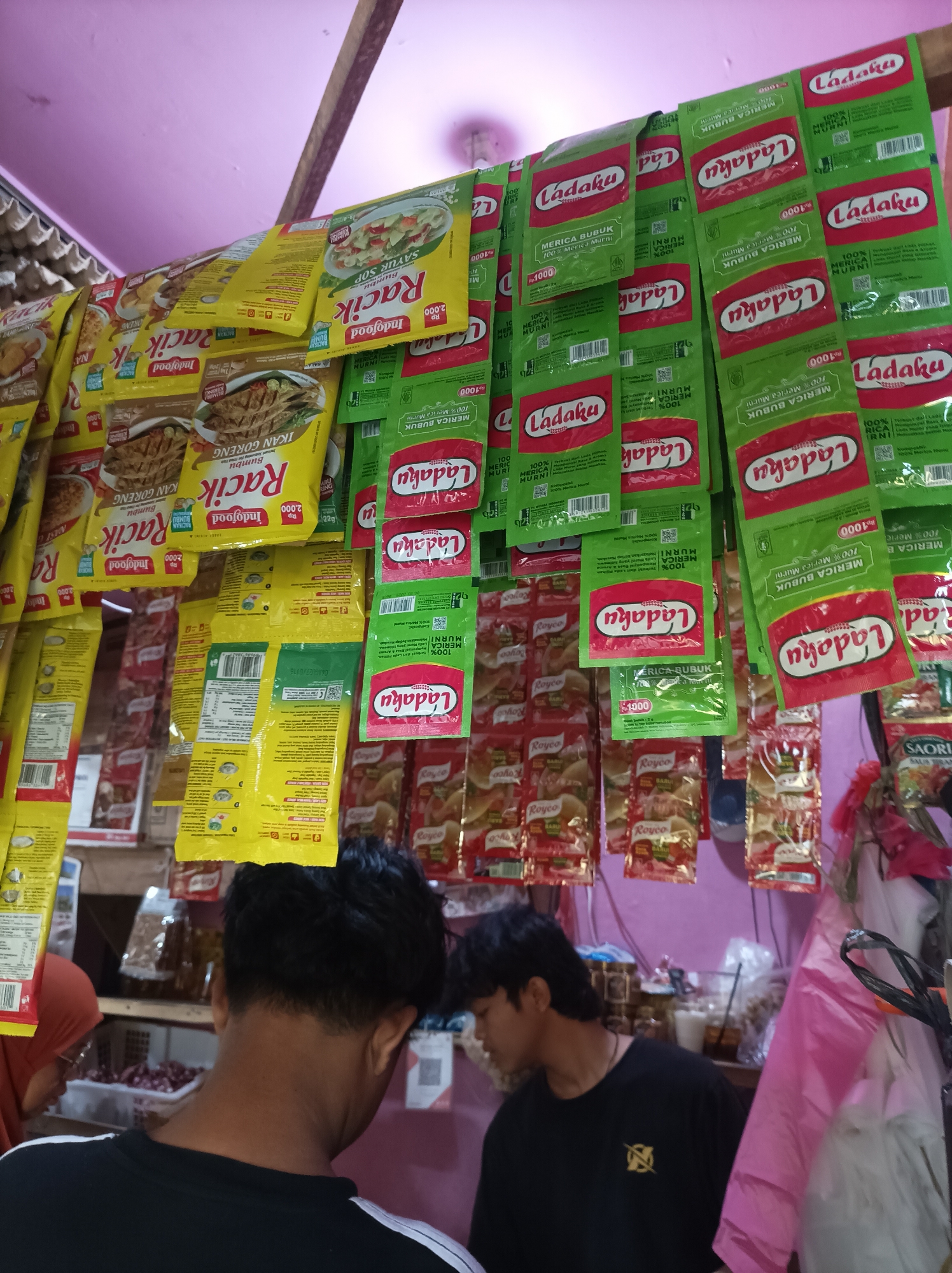 The image depicts a small, cluttered shop stall in West Jakarta, Indonesia, likely a warung or minimarket. The stall is overflowing with various packaged goods, primarily spices and seasoning mixes. Prominent among these are numerous packets of "Ladaku" brand pepper, displayed in green and red packaging. Yellow packets labeled "Racik" by Indofood, featuring images of fried fish and soup, are also visible.
The goods are hung from the ceiling and shelves, creating a dense display. Some of the packets are slightly blurred, suggesting movement or a shallow depth of field. In the foreground, the backs of two young men are visible. One, wearing a black t-shirt with white stripes on the sleeves, faces away from the camera. The other, also wearing a dark shirt, is partially visible to his right, looking towards the stall. The environment appears to be indoors, with a pink ceiling and a wooden structure overhead. The overall impression is one of a busy, local retail space with a wide variety of readily available household items.