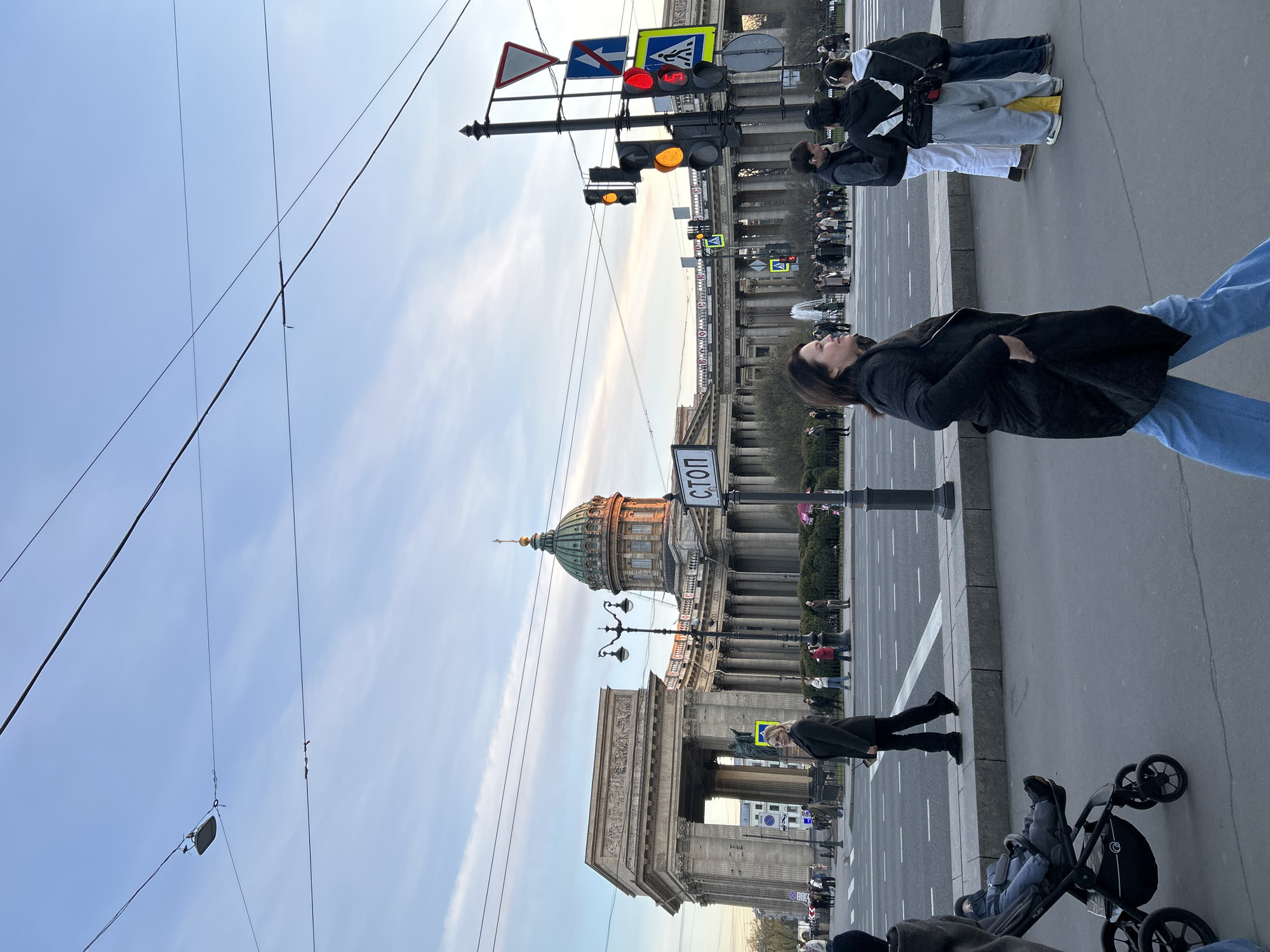 The image captures an urban street scene in Saint Petersburg, Russia, likely late afternoon or early evening, given the golden light on the buildings and the mix of blue and pale orange in the sky. The weather appears clear with scattered clouds.
In the foreground, a woman in a black jacket and blue jeans stands on the right, looking upwards. To her left, partially visible, are the legs of another person in dark pants next to a black baby stroller, in which a child is seated. Further back on the right, a group of people stand on a sidewalk, some wearing casual jackets and hoodies, one with a yellow bag.
The background is dominated by classical architecture. On the left, a grand building with columns and a distinctive green dome, characteristic of the Kazan Cathedral, is prominent, with sunlight illuminating its upper sections. Below it, an arched structure with detailed carvings is visible. A "СТОП" (STOP) sign in Russian is mounted on a pole near the cathedral.
On the right side of the street, multiple traffic lights are visible, some displaying red, others amber/yellow. A cluster of road signs, including a triangular yield sign, a blue square sign, and a pedestrian crossing sign, are attached to a pole above the traffic lights. Numerous overhead wires crisscross the blue sky, some with small insulators or connectors. Street lamps are also interspersed along the street. The road surface is asphalt with white painted lines for lanes and pedestrian crossings. People are seen walking or standing on sidewalks on both sides of the street, engaging in typical daily city activities. The overall scene depicts a busy historical area.