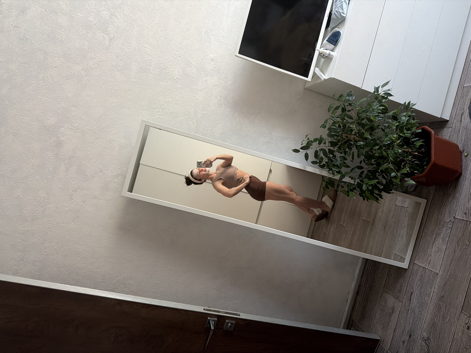 This image, rotated 90 degrees clockwise, captures an indoor scene, likely a hallway or room in Perm, Russia, featuring a woman taking a mirror selfie.
In the center, a tall, white-framed full-length mirror reflects a young woman. She is shown holding a silver-colored smartphone to her face, capturing her reflection. She has dark hair tied back with a white headband and is dressed in a light brown sleeveless top and dark brown athletic shorts, highlighting an athletic physique with defined legs.
To the right of the mirror, a vibrant green leafy plant in a brown rectangular pot rests on the light grey-brown wooden floorboards. Above the plant, a white wall-mounted cabinet or shelf unit is visible, with a blue and white shoe along with other items stored in one of its open compartments. The walls surrounding the mirror and cabinet are light-colored and textured, possibly finished with plaster or patterned wallpaper. In the lower left foreground, a dark wooden surface, which appears to be part of a door, is visible, equipped with a metal handle and lock mechanism.
The scene is well-lit, indicating a bright indoor environment, likely during the daytime. No discernible text is visible on any objects or surfaces within the image.