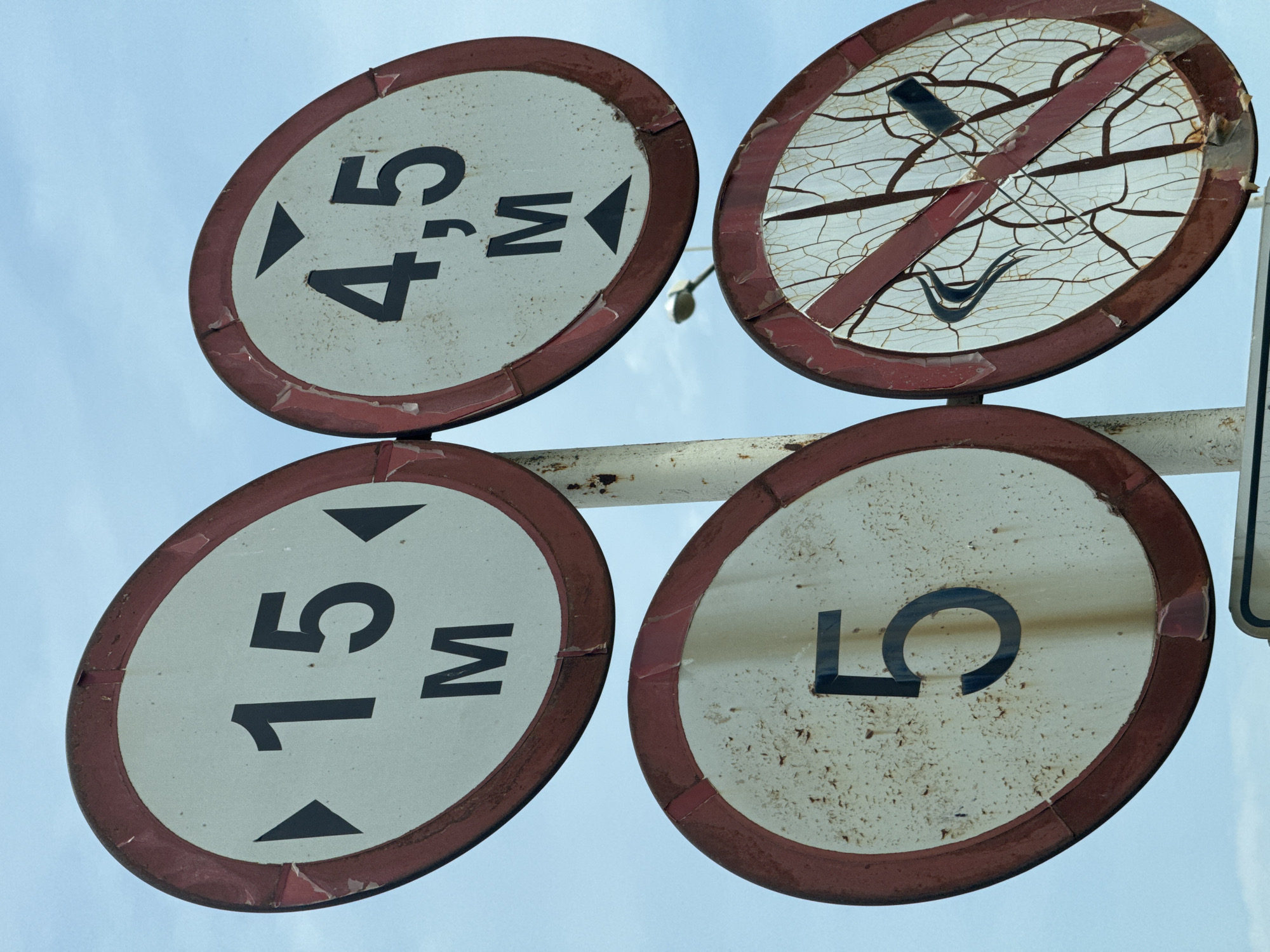 The image shows a collection of four weathered circular signs mounted on a white pole against a pale blue sky.
The top left sign is white with a red border. It displays the number "15" in large black font, with the letter "M" below it. Two black triangles point inwards from the left and right sides of the number "15". This sign indicates a maximum speed limit or a measurement.
The top right sign is similar in design, white with a red border. It shows "4,5" in black numbers, with a comma separating the digits, and "M" below. A black triangle points downwards from the top of the sign, and another points upwards below the "M". This sign likely indicates a maximum height restriction of 4.5 meters.
The bottom left sign is also white with a red border and features a large black numeral "5". This sign probably represents a speed limit of 5.
The bottom right sign is highly damaged, with a white surface covered in a network of fine cracks and peeling paint. It has a red diagonal stripe across it and a symbol of a lit cigarette with smoke, also crossed out by the red stripe. This is a "No Smoking" sign.
The signs are old and show signs of rust and wear, particularly around the edges and on the surfaces. The sky is clear and slightly overcast. In the background, a street light can be seen. The context suggests these are traffic or regulatory signs, possibly located in an area with restrictions on vehicle height and speed, and a prohibition on smoking. The image is framed from a low angle, looking up at the signs.