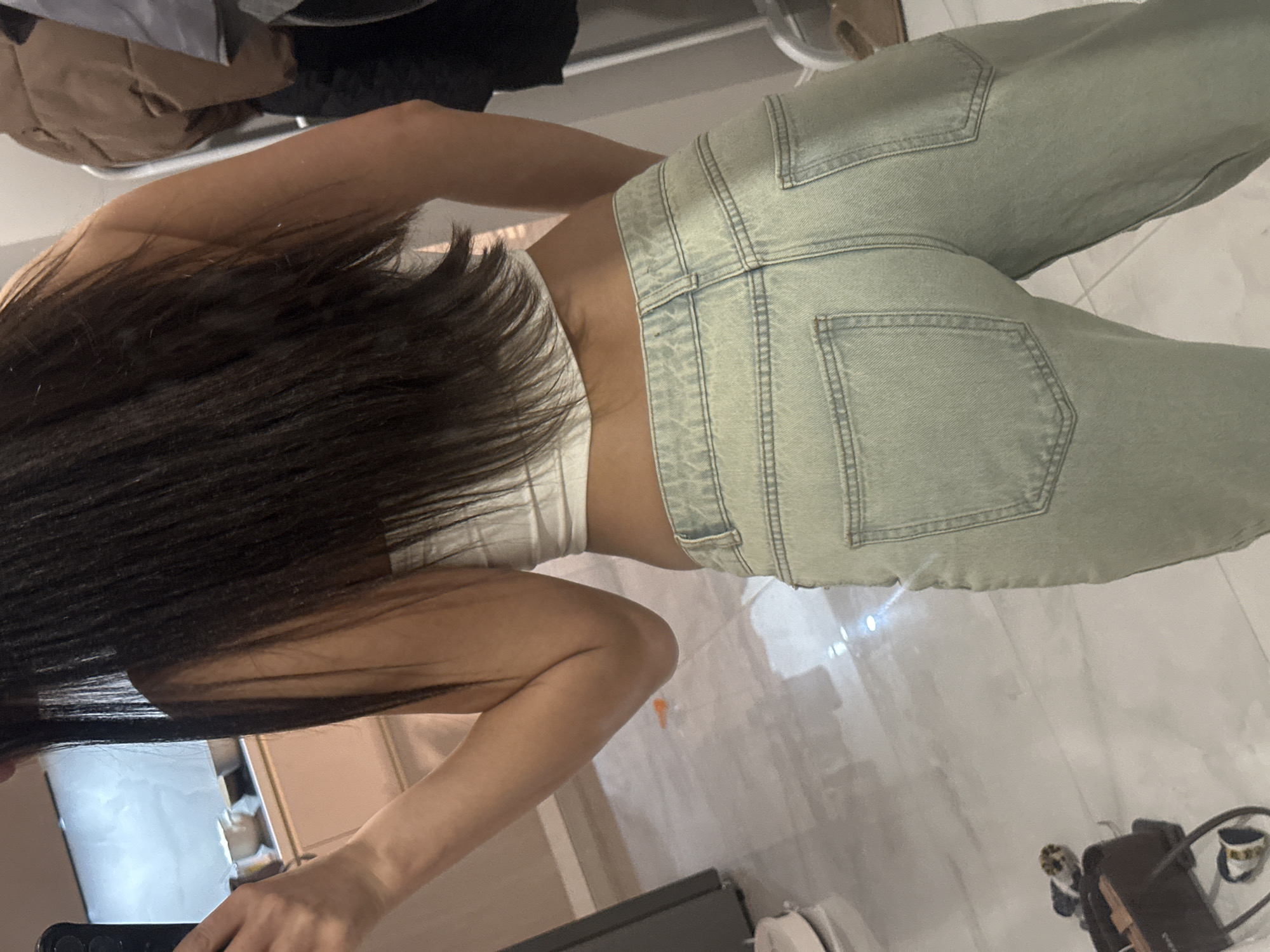 A person with long, dark hair is standing indoors, facing a mirror, wearing a white crop top and light green denim jeans. The person is holding a smartphone in their left hand, presumably to take the picture. The environment includes a tiled floor and possibly a bathroom counter. There is no information available to determine the location of the image in Khimki, Russia.