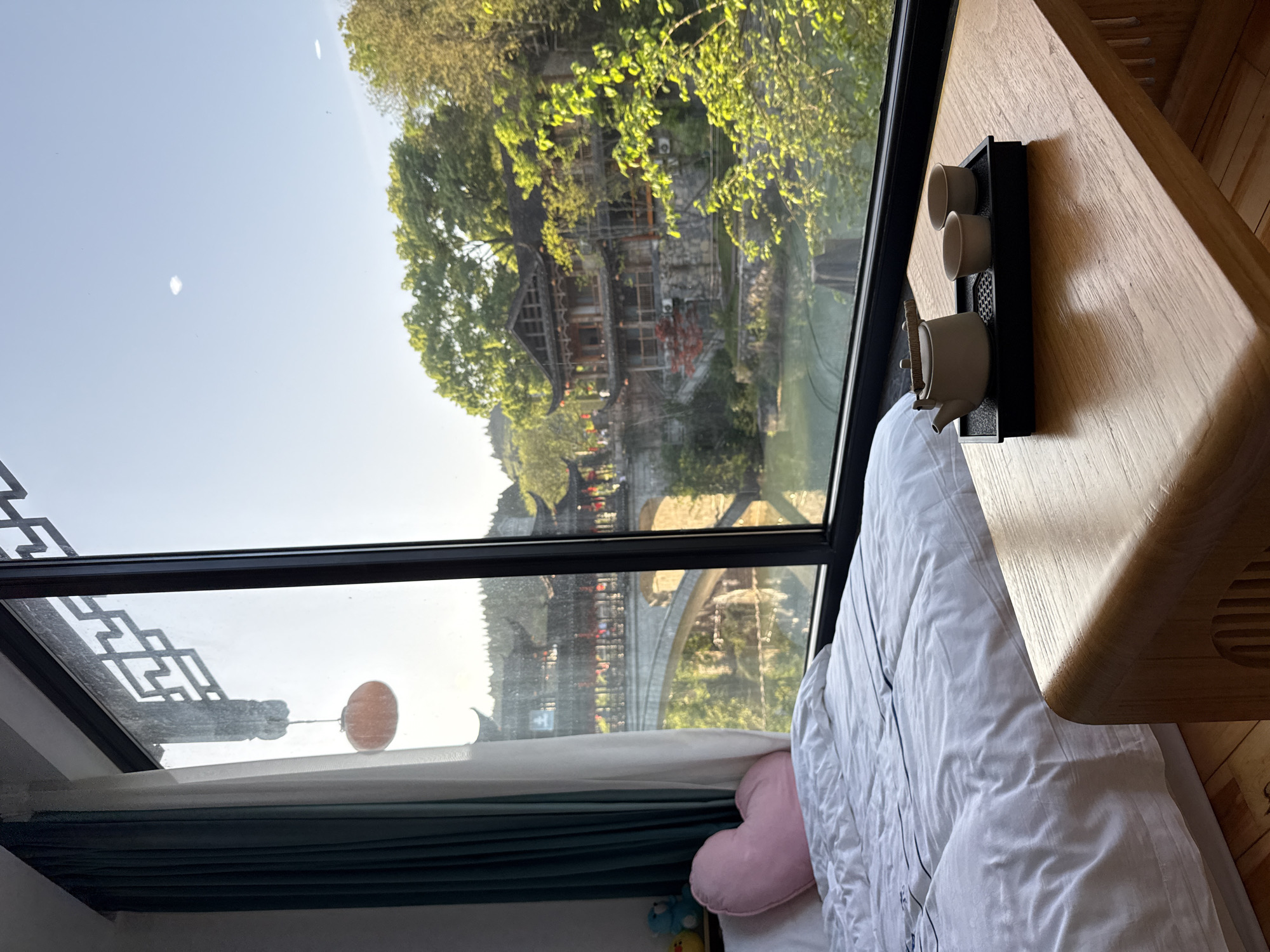 The image depicts a view from a bedroom or lodging in Furong, China. The window, with a decorative top frame, provides a view of the city. Traditional buildings and bridges are visible through the window. A tea set sits on a wooden table positioned near the window. A white comforter covers the bed. A pink heart-shaped pillow rests on the bed against a green curtain.