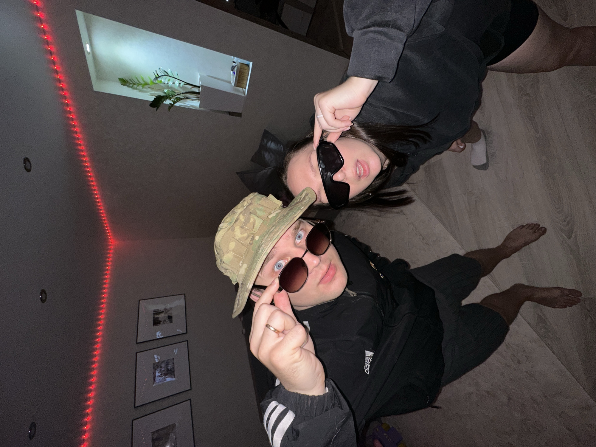 This indoor photograph, taken in Chaikovsky, Russia, features two individuals posing for the camera. The image is oriented such that the floor appears to be on the right side and the wall on the left.
In the foreground, a person (left) wears a camouflage bucket hat, dark sunglasses, and a black track jacket with the "Adidas" logo visible on the right side of the chest. They are also wearing dark shorts and are barefoot. This individual is looking directly at the camera, with their left hand lifting their sunglasses slightly, revealing blue eyes. They wear a simple band on their ring finger.
Behind and slightly to the right, another person (right) is visible. They are wearing a dark long-sleeved top and dark shorts, with one foot visible in a white sock. This individual also wears dark, rectangular sunglasses and is looking towards the camera, slightly pulling down their sunglasses with their right hand.
The setting appears to be a modern room with wooden or laminate flooring and light-colored walls. A string of red LED lights runs vertically along the wall on the left side of the frame. Below these lights, a stack of three framed pictures is visible. Further up the wall, there's a recessed alcove containing a green plant, illuminated by an upward-facing light. The overall lighting suggests an evening or dimly lit indoor environment, relying on artificial light sources. Both subjects exhibit a casual and direct demeanor.