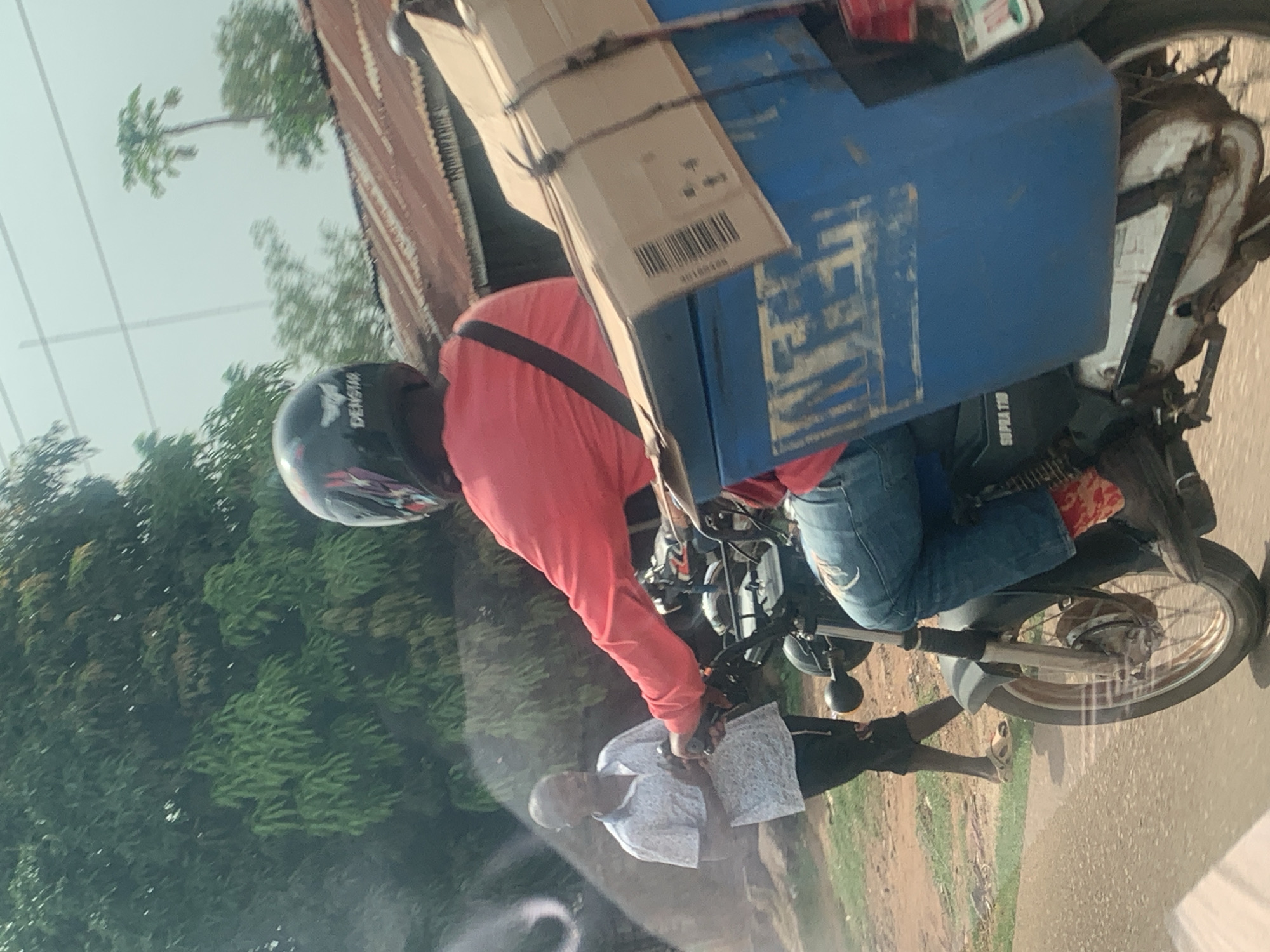 The image captures a scene in Maigizo, Nigeria. A motorcycle rider wearing a black helmet and red long-sleeved shirt is transporting a large blue container and a cardboard box secured on the bike's rear. The rider is wearing blue jeans. Adjacent to the road, a man in a patterned white shirt and dark shorts stands on the grassy shoulder, near the motorcycle. The surrounding environment includes lush green trees. A building with a rusted roof is partially visible. The motorcycle has a license plate and "SUPRA T100" written on it.