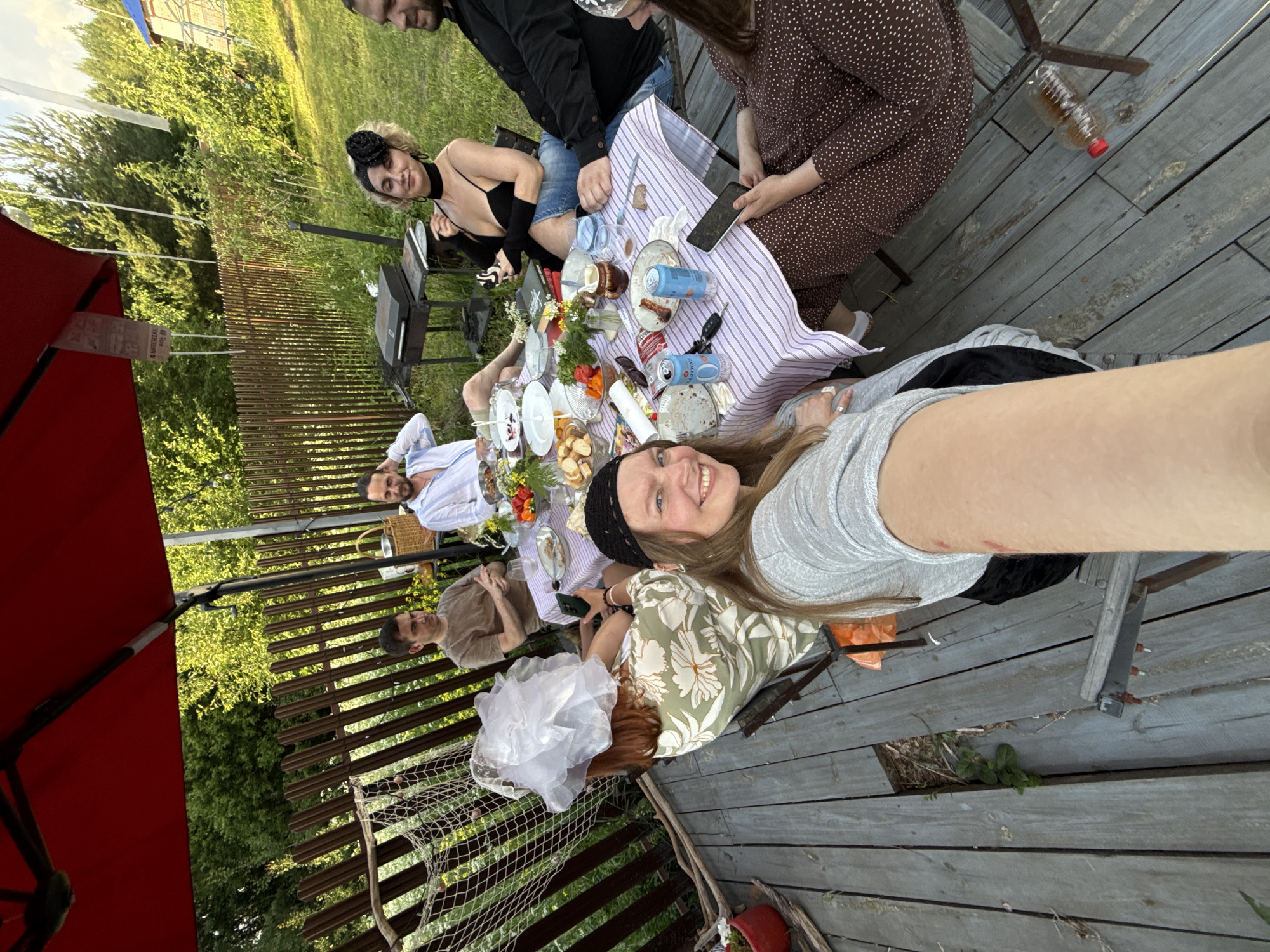 The image captures an outdoor gathering of approximately seven individuals seated around a table laden with food and drinks on a wooden deck. The setting appears to be a backyard in Izhevsk, Russia, with a wooden fence and lush green foliage visible in the background. A large red umbrella provides shade.
The people are dressed casually, with one woman in the foreground holding a phone and appearing to take a selfie. She is wearing a grey top and a black skirt. To her left, another woman is visible wearing a floral print dress and a large white fascinator. Further around the table, a man in a brown t-shirt is seated, and next to him, a man in a white shirt is seen. Another woman, sporting a black headpiece and a black top, is seated opposite the selfie-taker. A man in a black shirt is partially visible on the right side of the frame.
The table is covered with a striped tablecloth and features plates with food, bottles, cans, and glasses. Some of the food items appear to be snacks and appetizers. A grill is situated behind the table, suggesting a barbecue event. The overall atmosphere suggests a relaxed social gathering.