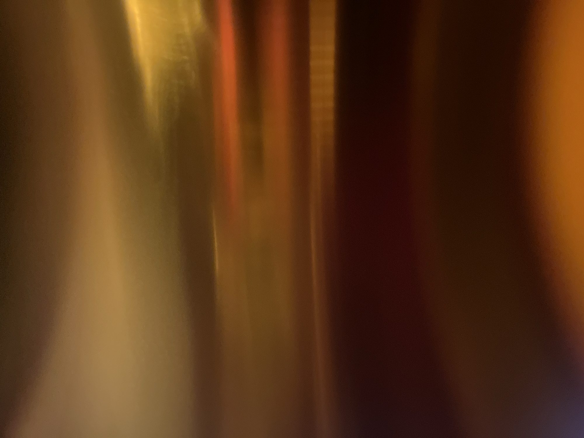 The image displays an abstract, out-of-focus, horizontal composition of warm colors, primarily shades of orange, yellow, and brown. There are no discernible objects, people, or specific locations identifiable within the frame. The lighting appears to be artificial, creating blurred streaks and gradients of light and shadow. The overall impression is one of atmospheric abstraction rather than a representational scene.