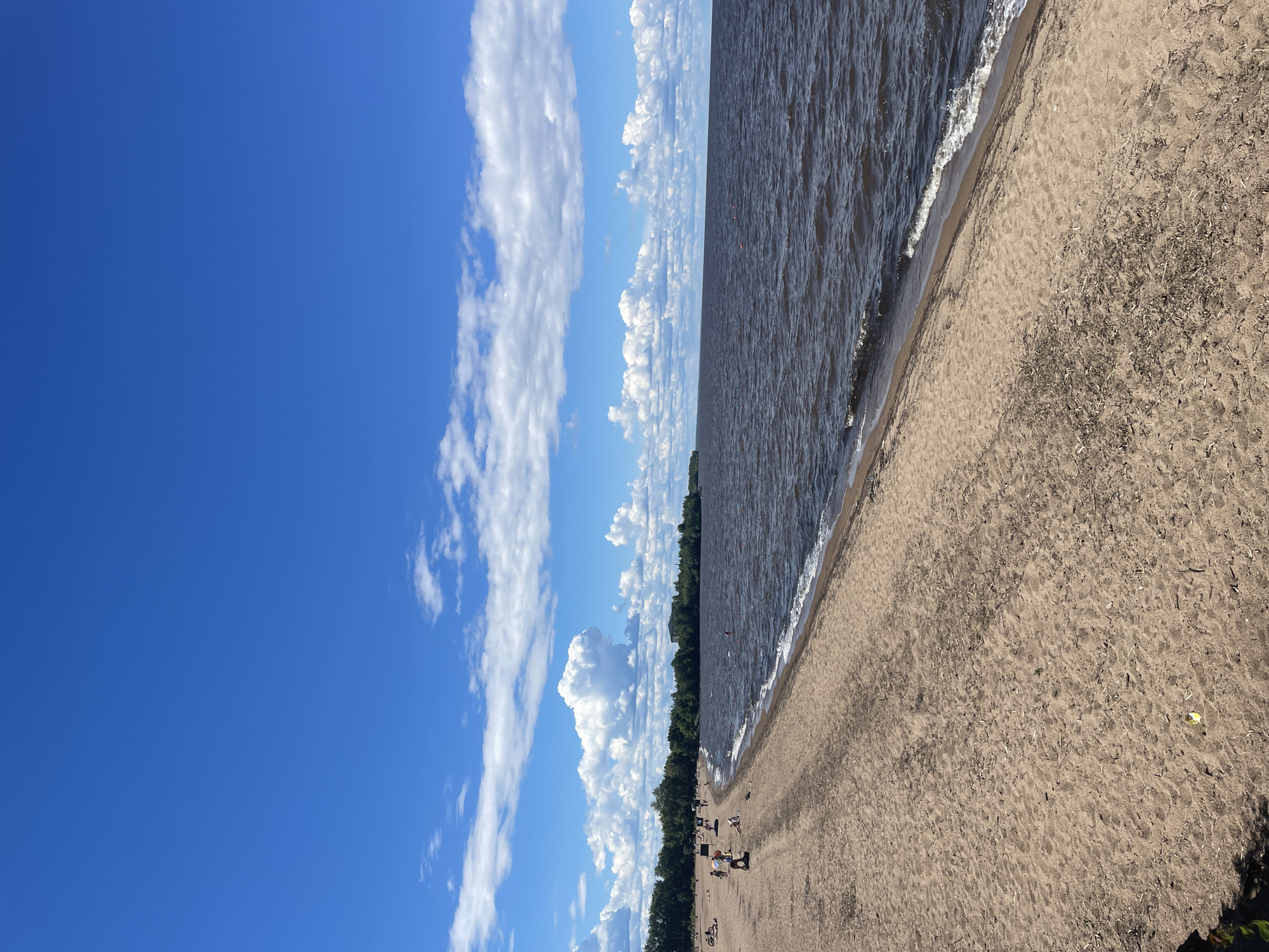 The image shows a sandy beach on the shore of a large body of water, likely Lake Beloye, in Belozersk, Russia. The sky is bright blue with scattered white cumulus clouds. The water is dark and choppy with small waves breaking on the shore. A line of green trees borders the beach in the distance. There are several people on the beach, some sunbathing, others walking or sitting. A few people are in the water. The sand is light brown and appears to be a mixture of sand and small debris. The overall scene depicts a warm, sunny day at the beach.