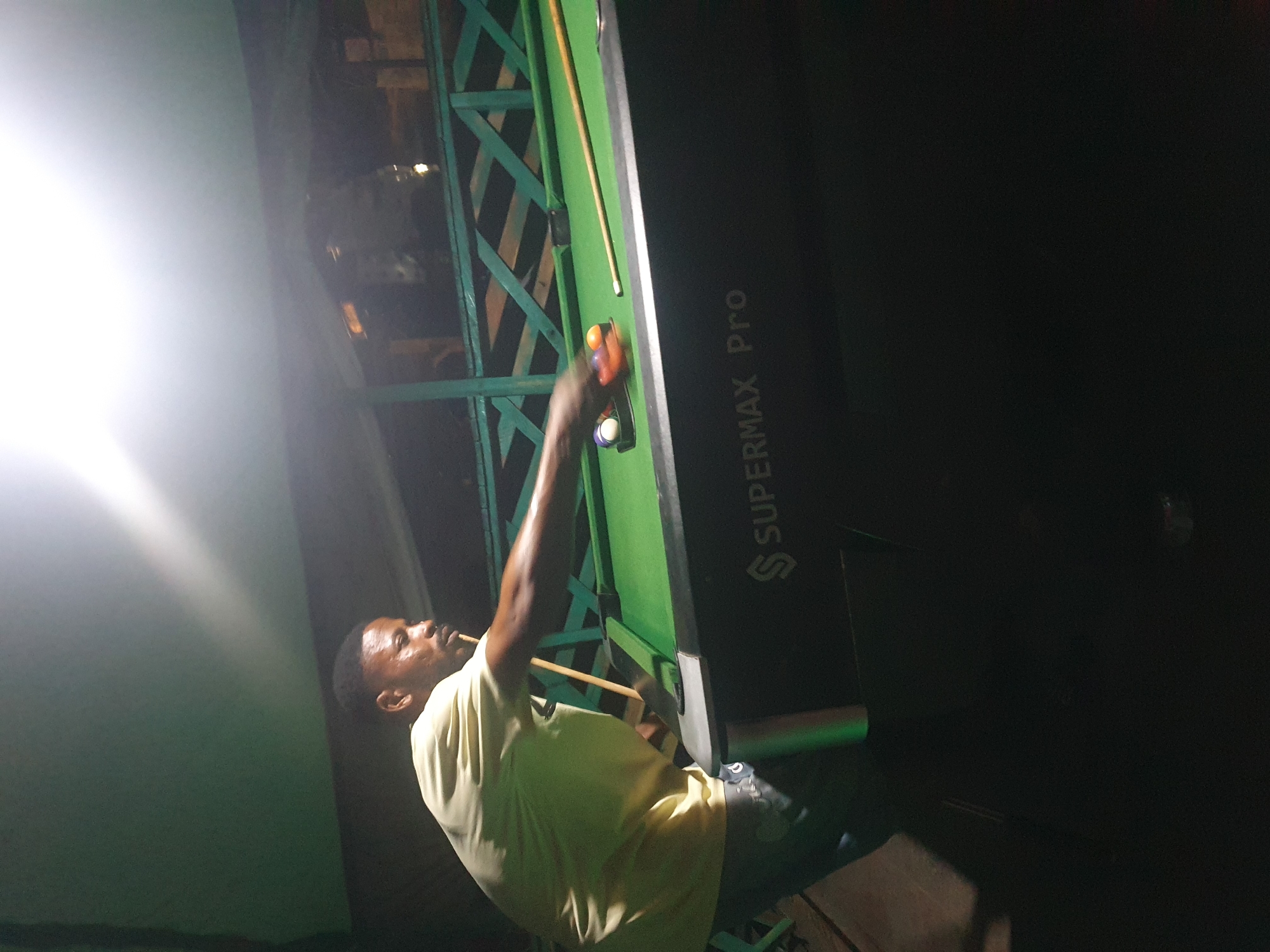 A medium shot captures a Black male with short hair and a mustache playing pool. He is wearing a light yellow t-shirt and dark pants. He stands on the left side of the image, facing right, with his body angled towards a green pool table. His right arm is extended, holding a pool cue aimed at a cluster of pool balls on the table. The pool table has the brand name "SUPERMAX Pro" visible on its side. The environment appears to be an outdoor or semi-outdoor setting with a dark, possibly wooden, railing in the background and a green ceiling or awning overhead, brightly lit by an overhead light source. The image was taken at night in Lokoja, Nigeria.