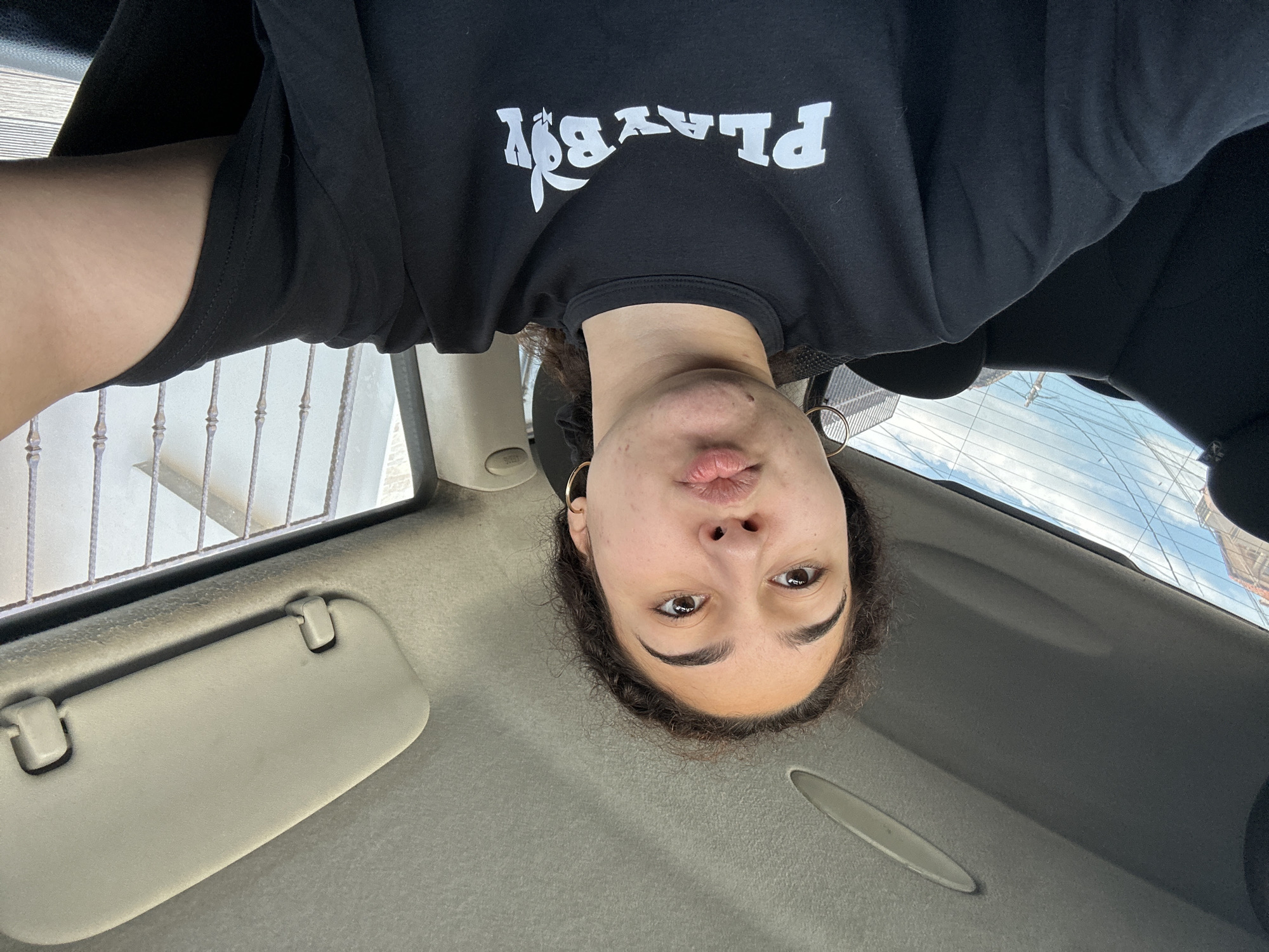 A selfie-style photograph presents a person inside a vehicle, possibly a car. The photograph is taken from a slightly upside-down perspective.
The person is wearing a black t-shirt with a white "Playboy" logo. The focus is on the person's face, with visible facial features.
The interior of the vehicle includes a sun visor and the roof. Through the window, a view of the outside world is visible, showing buildings and the sky.
The environment suggests the location may be urban, although specific details connecting to Burgas, Bulgaria are not immediately apparent in the photo. Without more visible landmarks, architectural styles, or clear signage, a precise location within the city is not discernible.