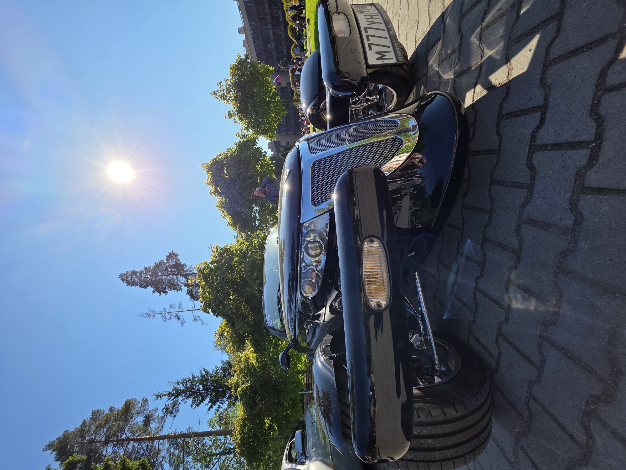 A close-up, vertically oriented photograph shows the front of a black vehicle parked on a paved surface in what appears to be a parking area or roadside setting. The vehicle has a distinctive chrome grill and headlights. The Russian license plate "M777YH" is visible on another black vehicle slightly behind the first. Trees and buildings are visible in the background under a bright blue sky with the sun shining, indicating it is daytime. People can be seen in the background near the buildings. The location is indicated to be in Repino, Russia.