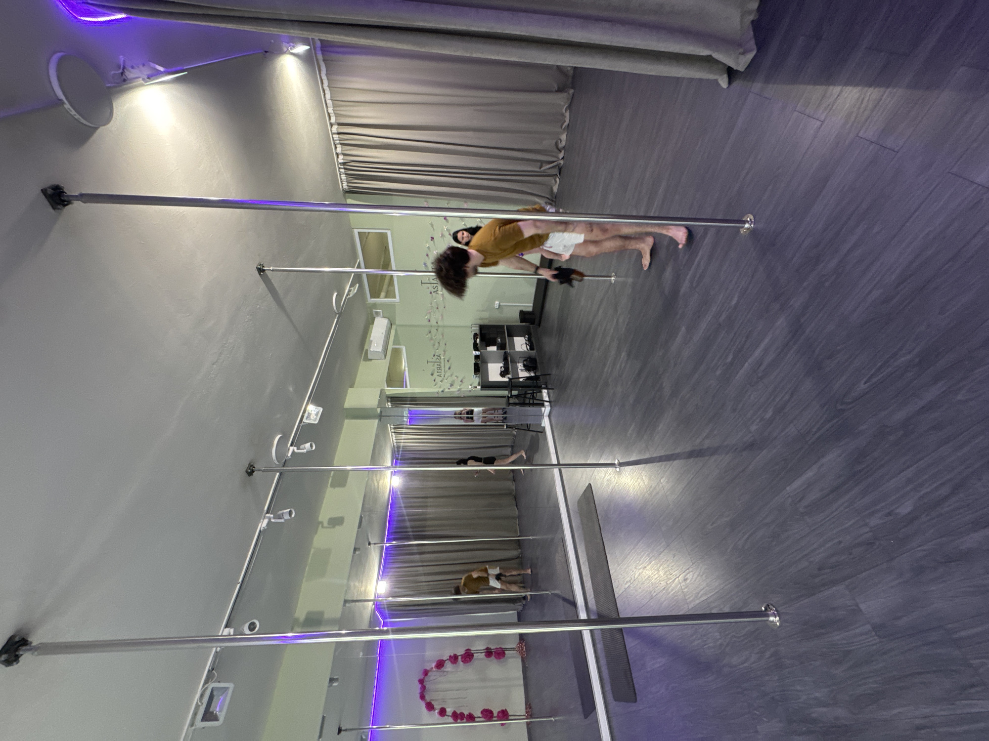 Here is a description of the image:
The image shows an interior view of a fitness studio with pole dancing equipment. The studio has multiple vertical poles anchored to the ceiling and floor, arranged in a linear fashion. The walls are painted in a neutral tone, likely gray or light beige. Large curtains adorn one of the walls. The flooring appears to be a dark, wood-look material.
In the foreground, a person wearing an orange shirt and shorts is positioned horizontally on one of the poles. There are other individuals performing activities on different poles in the background.
The studio is well-lit with a combination of natural and artificial lighting. Some of the lighting fixtures emit a purple or blue hue. The composition is somewhat distorted due to the camera angle, but the overall setting is clear as a fitness studio designed for pole dancing.
