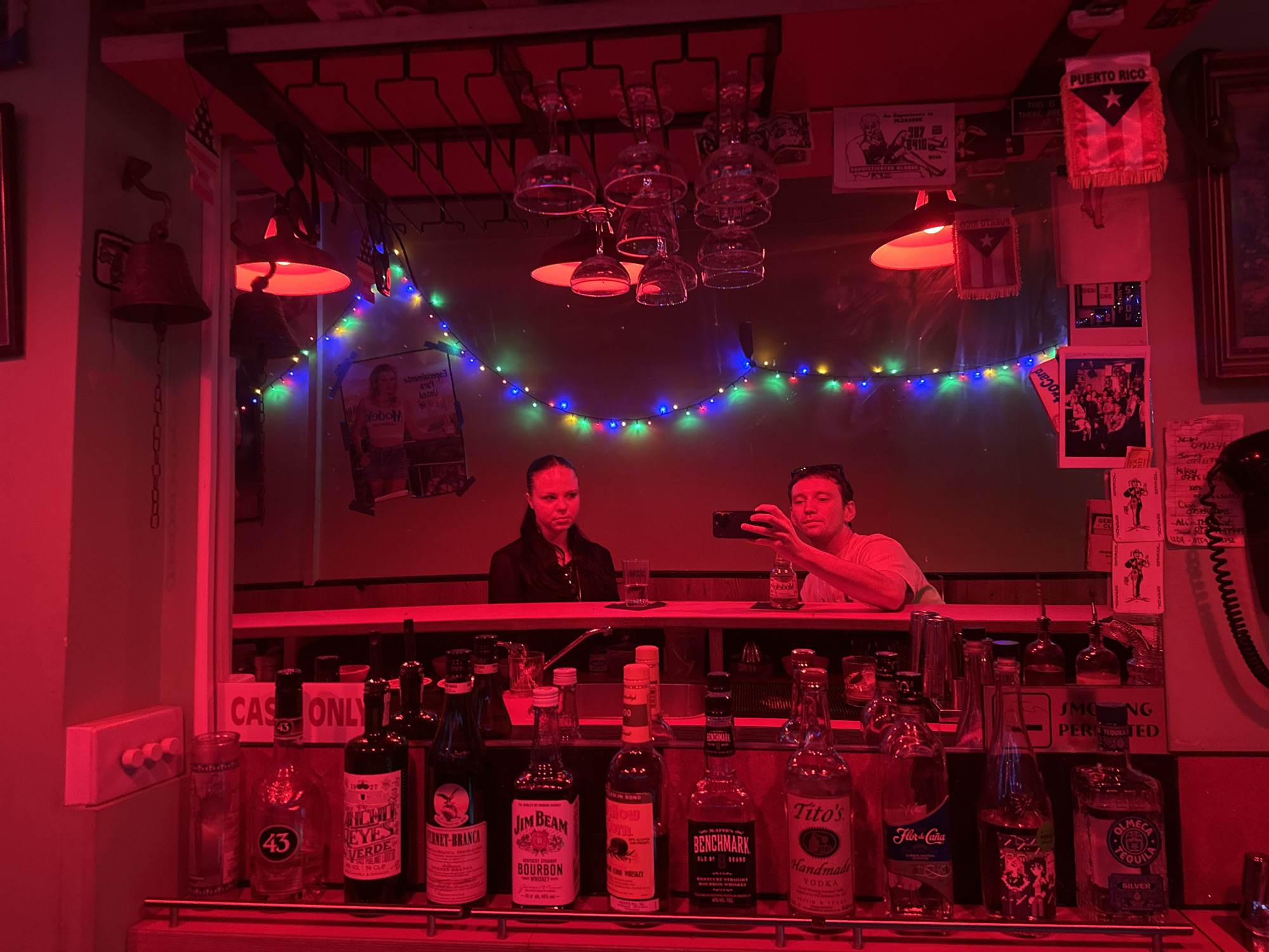 The image shows the interior of a bar, likely in Puerto Rico, based on the Puerto Rican flags and signage. The bar is dimly lit with red lighting and colorful string lights. There are two patrons visible behind the bar; a woman is looking straight ahead, and a man is holding a phone, possibly taking a photo. The bar counter is stocked with various liquor bottles, including brands like Jim Beam, Tito's, and Fernet-Branca. There are also signs indicating "Cash Only" and "No Smoking Permitted". Wine glasses are hanging upside down from a rack above the bar. Additional posters and decorations adorn the walls.