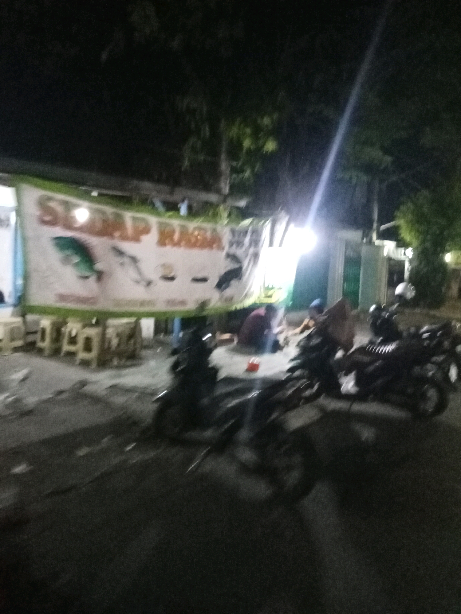 An out-of-focus, nighttime outdoor scene in Kepanjen, Indonesia, captures a street-side establishment with a banner displaying stylized images of fish. The banner, with green text that appears to read "SAMP RASA," is hung above a row of small tables and chairs. Several motorcycles are parked on the asphalt in front of the establishment. Three individuals are visible sitting on the ground near the tables, with one appearing to wear a blue head covering. The lighting is dim, with a bright light source to the left casting reflections and a shaft of light extending diagonally across the image from the upper right. The environment suggests a casual, open-air dining or gathering space.