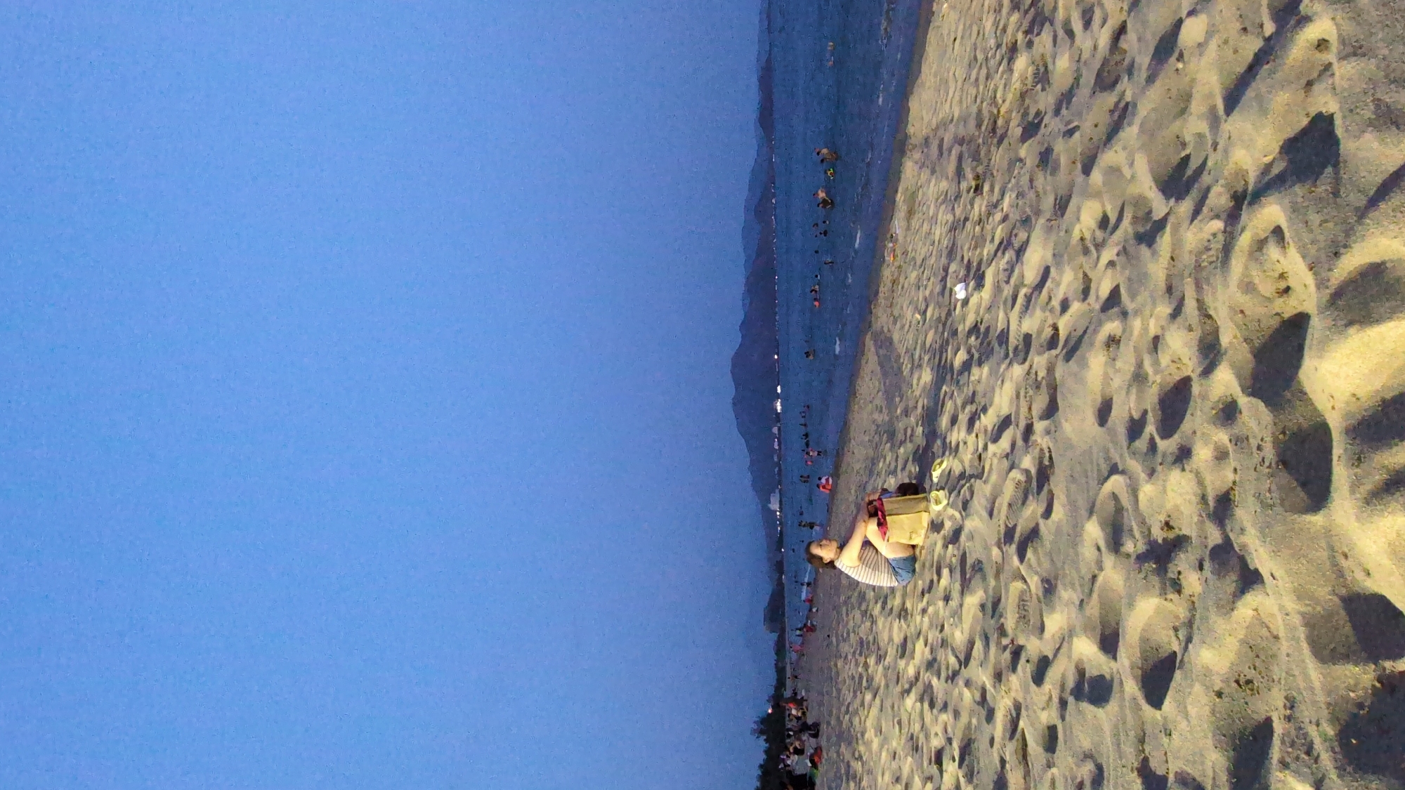 The image shows a beach scene, presumably in Khánh Hải, Vietnam, at dusk or twilight. The sky is a gradient of deep blue to lighter blue near the horizon. The beach is sandy, with numerous footprints visible. People are sparsely scattered along the shoreline and in the water. A person is seated on the sand near a box. The background contains a mountain range.