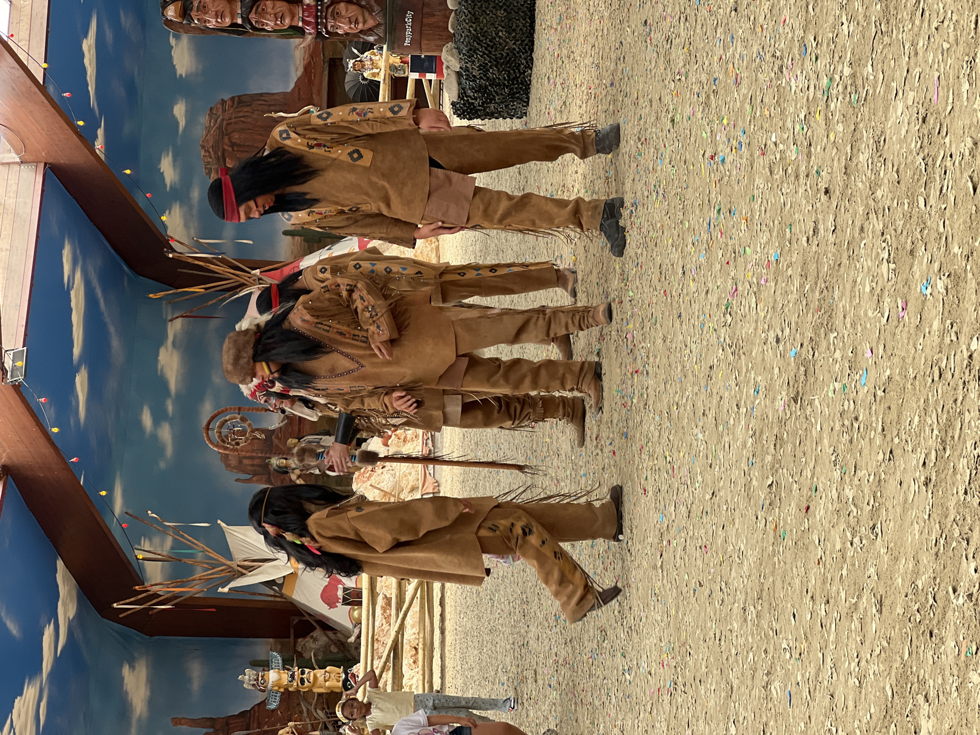 This image depicts an outdoor performance with individuals dressed as Native Americans. The scene appears to be set in a themed park or attraction, possibly related to a historical or cultural reenactment. In the foreground, three people in traditional Native American attire are the primary subjects. They are wearing brown leather outfits with fringes and decorative patterns. The person on the left is a child, facing left and walking away from the camera. The two individuals to their right are adults. One adult is facing forward and slightly to the left, while the other is facing to the right and looking down. A fourth person, also in Native American attire, is partially visible behind the first adult, facing left.
The background features a painted mural of a desert landscape with mesas and a cloudy sky. There are also several props, including teepees, totem poles, and a dreamcatcher. A wooden fence runs across the middle ground. In the far background, a structure with a wooden roof and hanging lights is visible. The ground is covered in sand or dirt, with scattered confetti. On the right side of the image, a wooden barrel with the text "PonyParkCity" is visible, suggesting the location is PonyParkCity in Collendoorn, Netherlands. Other figures, including what appear to be mannequins or statues of Native Americans and a small statue of a horse with a rider, are also present in the background. The overall atmosphere suggests a lively event or attraction.