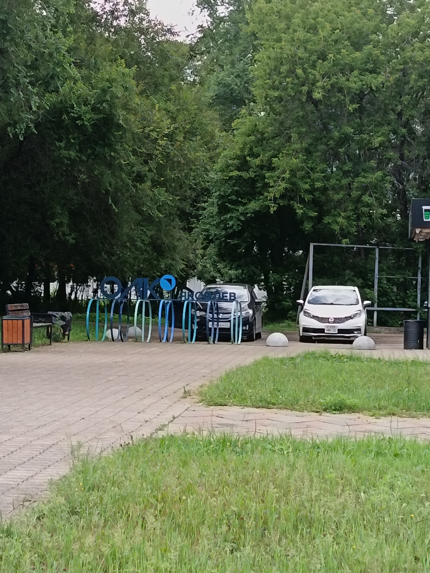 The image depicts a row of blue and green bicycle racks in front of a line of trees. To the right of the bicycle racks are two cars parked on a paved area. One is a dark-colored sedan with the letters "HEB" visible on its rear windshield. The second is a white hatchback with the license plate "CBZ 662". In the background, a large expanse of green trees forms a natural backdrop, suggesting a park or a wooded area within the city. The lighting indicates it is daytime.