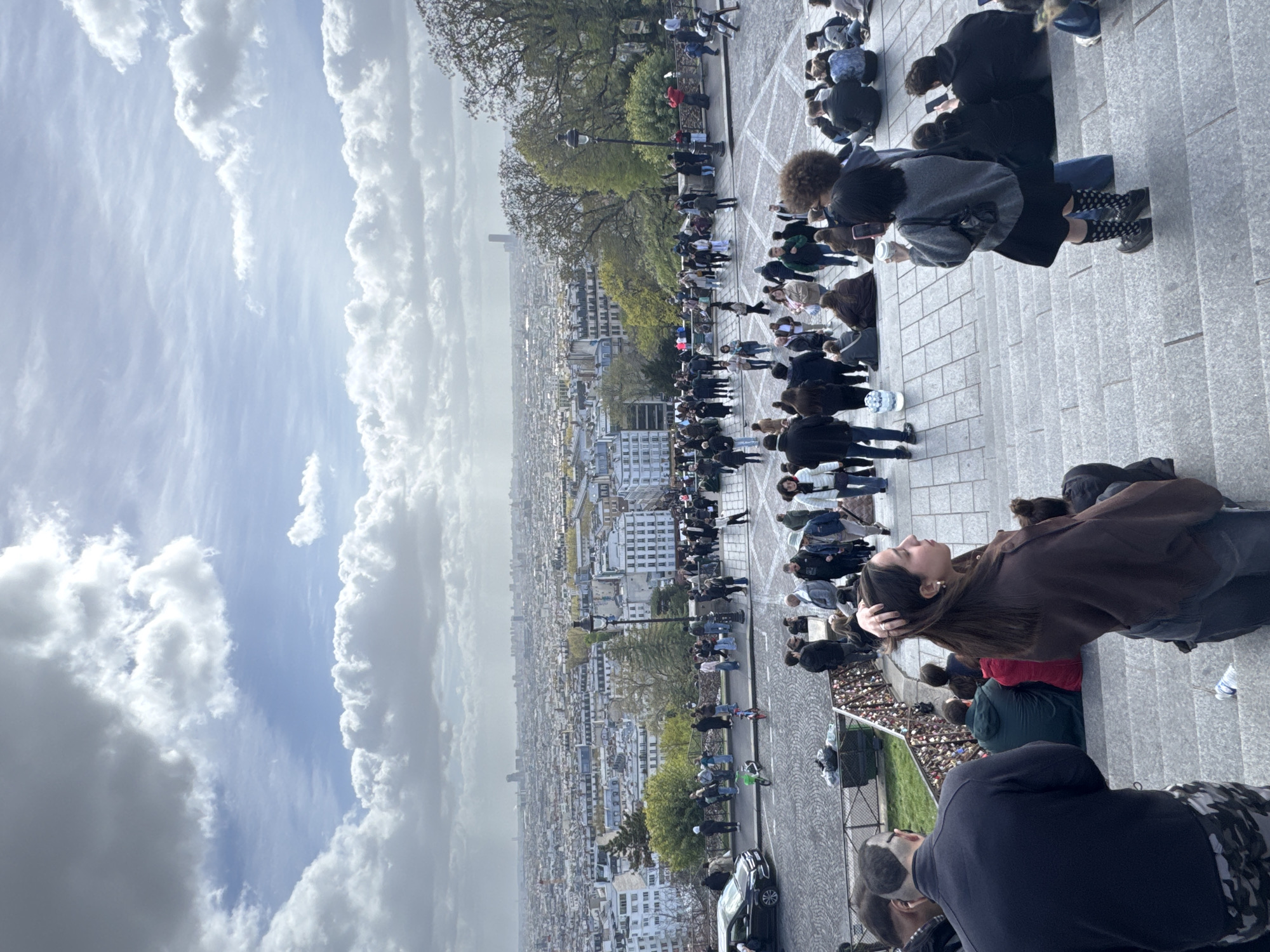 A long, medium-shot image shows a multitude of people, presumably tourists, congregated on the steps of the Sacré-Cœur Basilica in Paris, France. The Basilica is not visible, but the steps and the city view below give away the location. The crowd is densely packed along the steps, with people standing, sitting, and taking pictures. Many are looking down at the sprawling city of Paris, visible in the background. The city is a vast expanse of buildings stretching to the horizon. The sky overhead is cloudy with patches of blue. People are dressed in a variety of casual clothes, indicating the season is likely spring or fall. There are also some lampposts and greenery visible, further suggesting the steps are on a public walkway or park area.