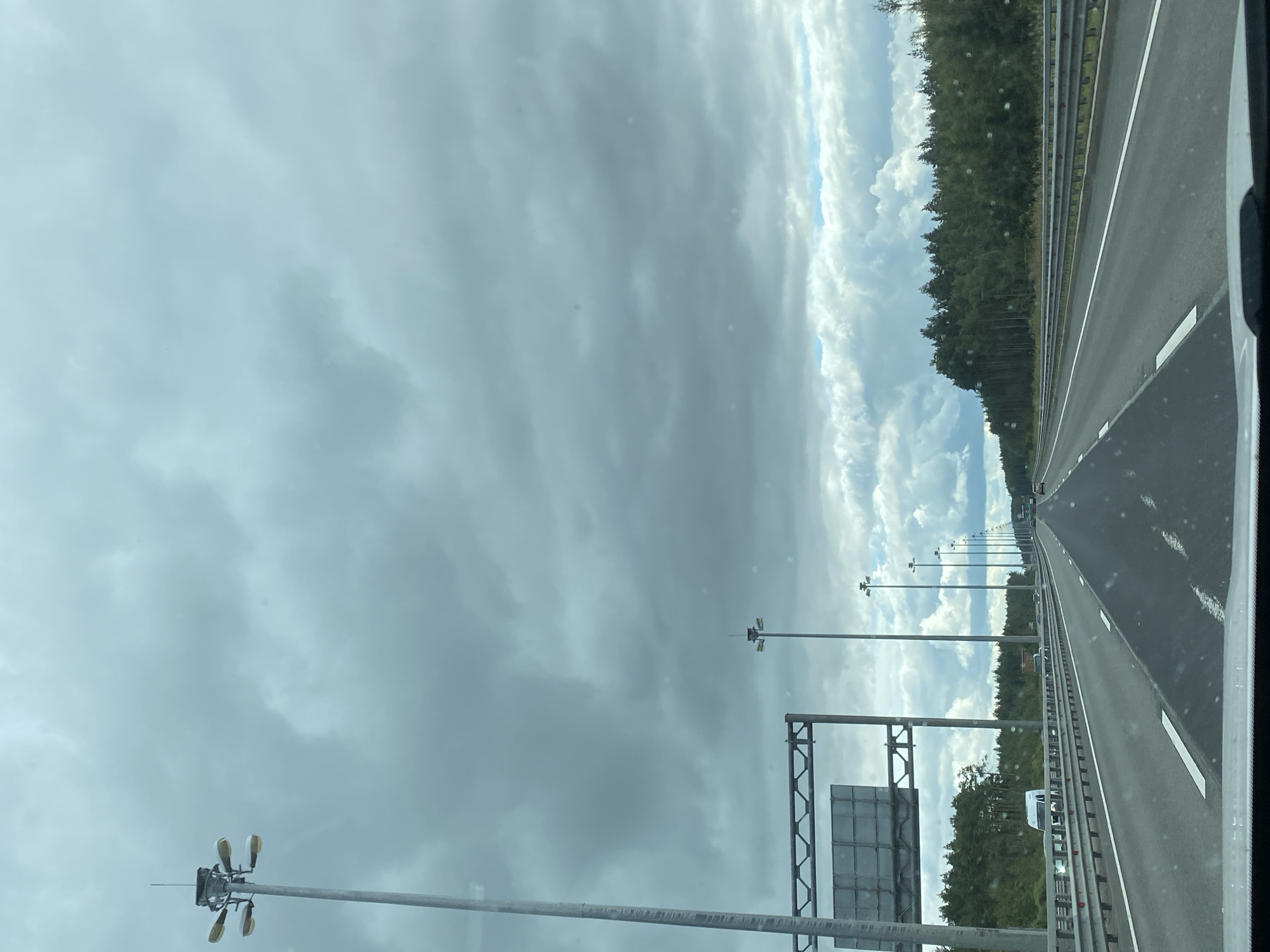 This image depicts a view from a vehicle traveling on a multi-lane highway, likely a federal or regional route, near Porzolovo, Russia. The sky is overcast with heavy, dark clouds, suggesting potential for rain or storm. Towering streetlights line the right side of the highway, with several visible in the distance. A large metal sign structure is present on the left side, with a blank rectangular sign. The highway itself has clear lane markings and is bordered by metal guardrails. On the left side of the highway, beyond the guardrail, a dense forest of pine trees runs along the road. A white van and other vehicles are visible in the distance on the highway. The perspective suggests the photograph was taken from the driver's or front passenger's seat of a car. The overall scene conveys a sense of travel on a major road under moody weather conditions.