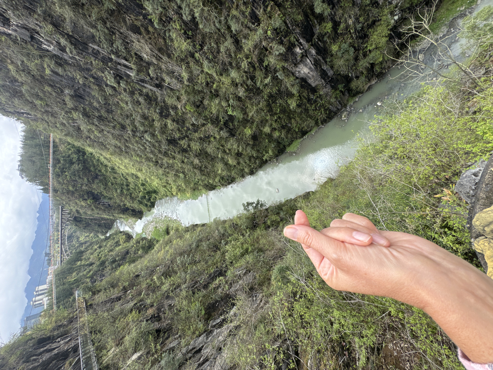 The image shows a view from a height looking down at a river running through a gorge in Qianjiang, China. Lush green vegetation covers the steep slopes of the gorge. In the foreground, a hand is making a "finger heart" gesture. In the distance, there is a bridge and some buildings, suggesting a developed area nearby. The river's water appears pale green.