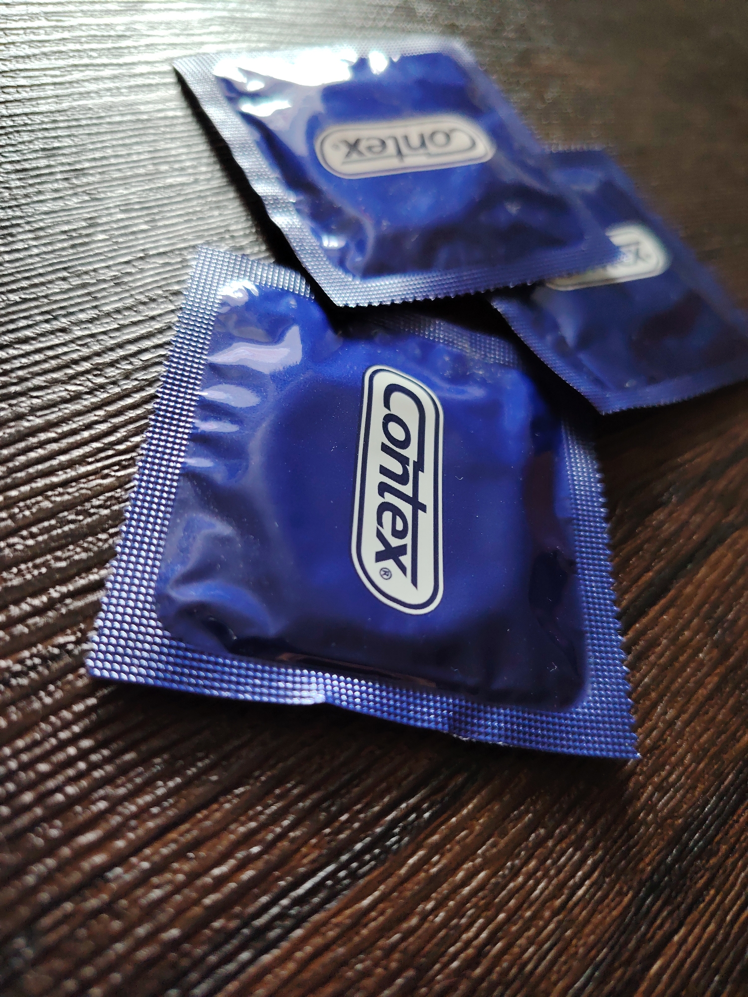 The image shows three blue Contex condom packets on a dark brown wooden surface. The packets are arranged haphazardly, with one packet fully visible in the foreground and two partially visible in the background. The lighting is somewhat subdued, creating shadows and highlighting the textures of the foil packaging and wood grain. The city of Chaikovsky, Russia, is not discernible from the provided image.