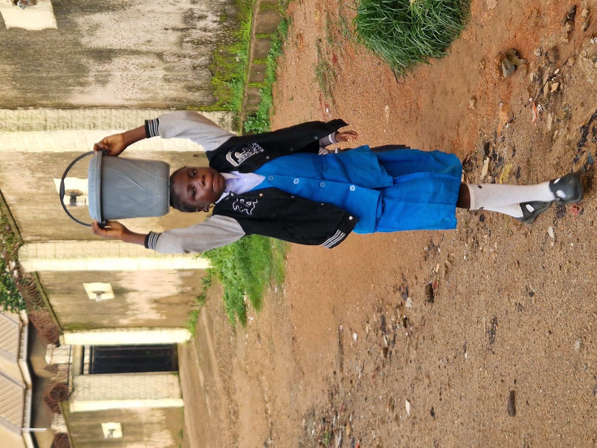 A young Black girl wearing a school uniform, a blue pleated skirt, a blue vest, and a gray jacket over a white shirt, walks on a dirt road in Kabong, Nigeria, carrying a large gray bucket of water on her head. She is facing the camera, with her arms raised to support the bucket. The road is unpaved and appears to be littered with debris. In the background, there is a concrete wall with a black metal gate. The environment appears to be an urban or semi-urban area with some vegetation.